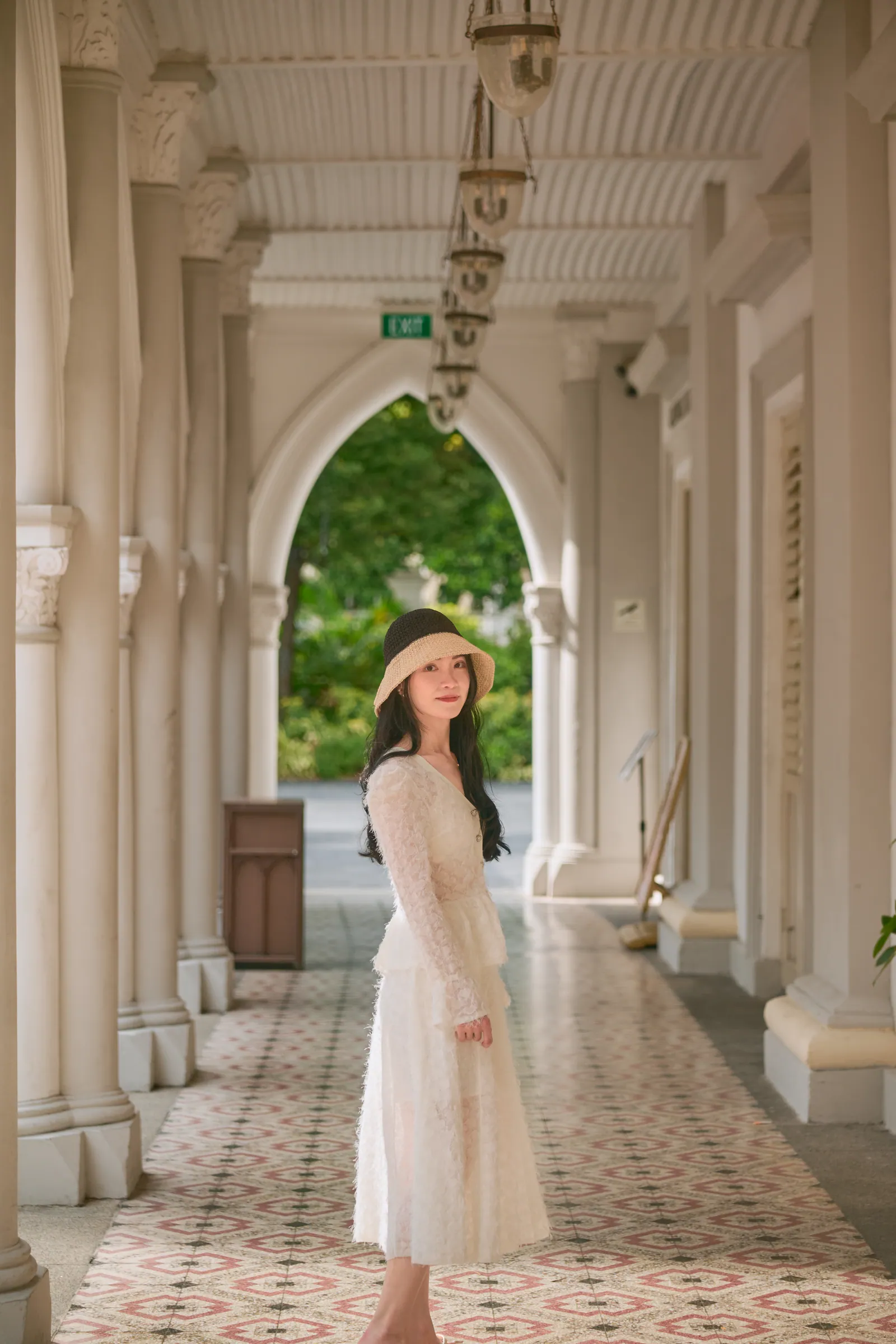 Woman in a cream lace dress and hat standing in a white arched corridor with patterned floor tiles, CHIJMES, Singapore