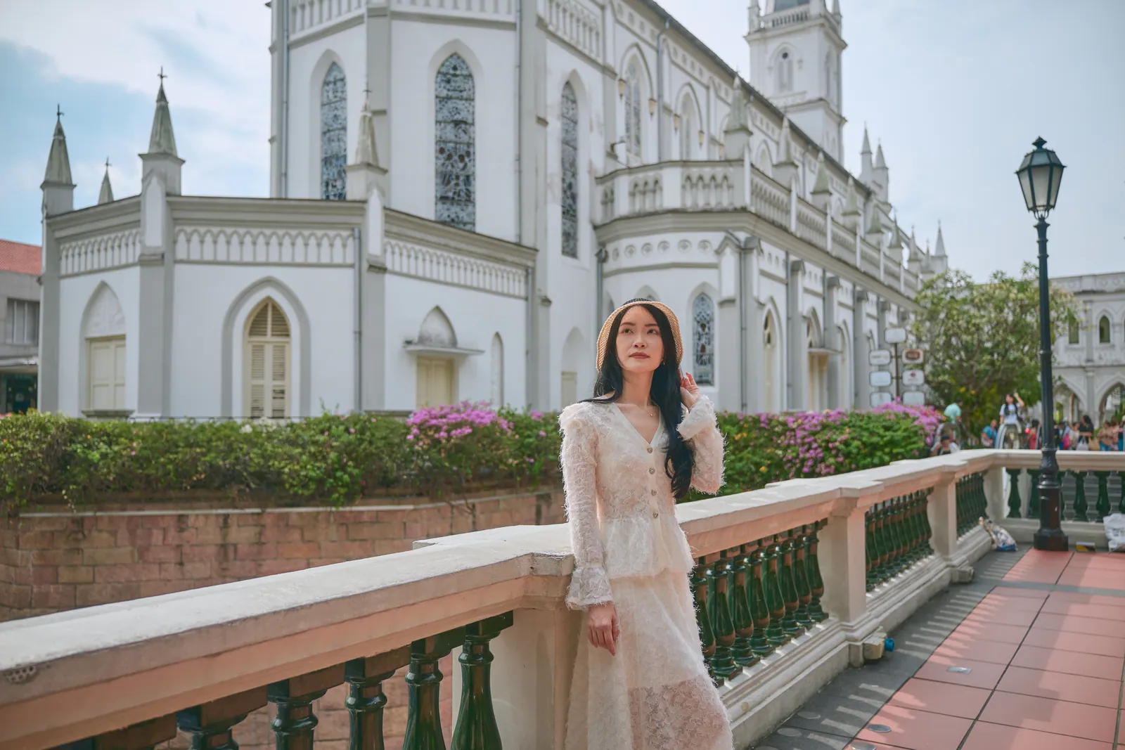 Woman in a cream lace outfit leaning on a white balustrade beside the Gothic chapel and pink flowers, CHIJMES