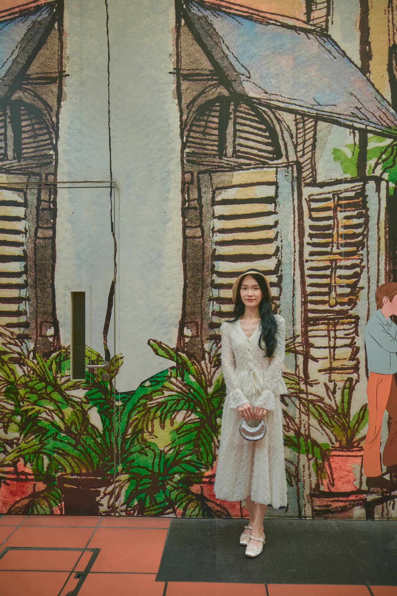 Woman in a cream lace dress standing in front of a painted mural of old shophouses and plants, CHIJMES