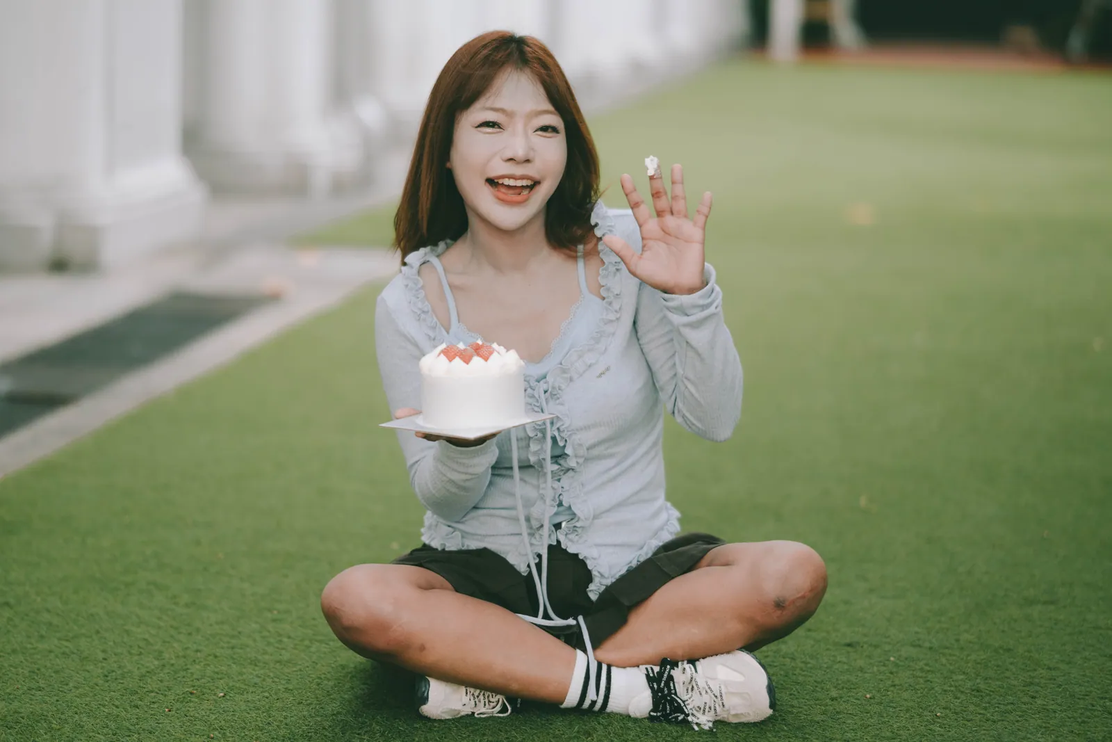 Woman sitting on a lawn with her strawberry birthday cake, laughing and waving, Singapore birthday photoshoot
