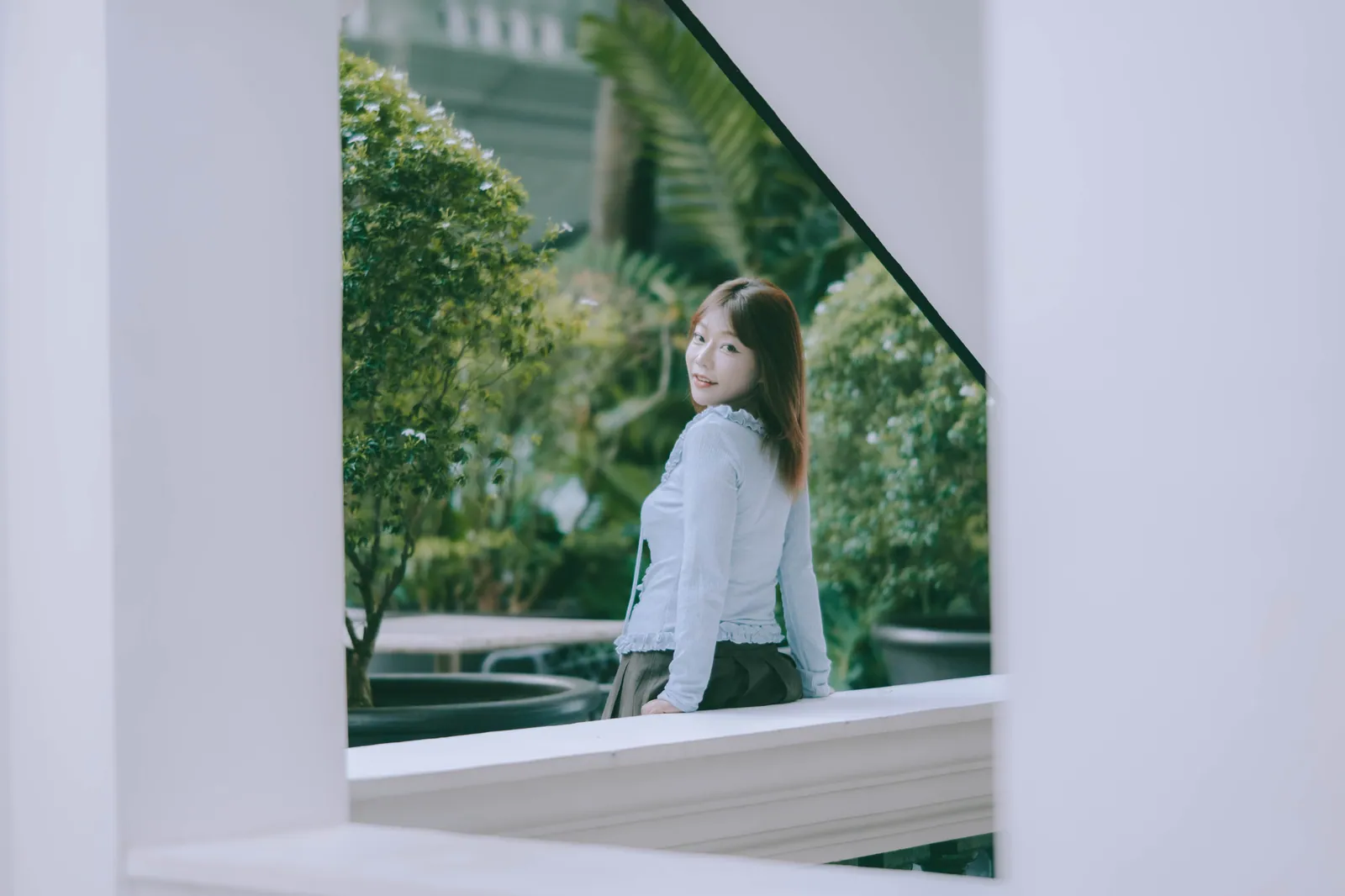 Woman in a pale blue ruffled top looking back over her shoulder, framed by white walls and greenery in Singapore