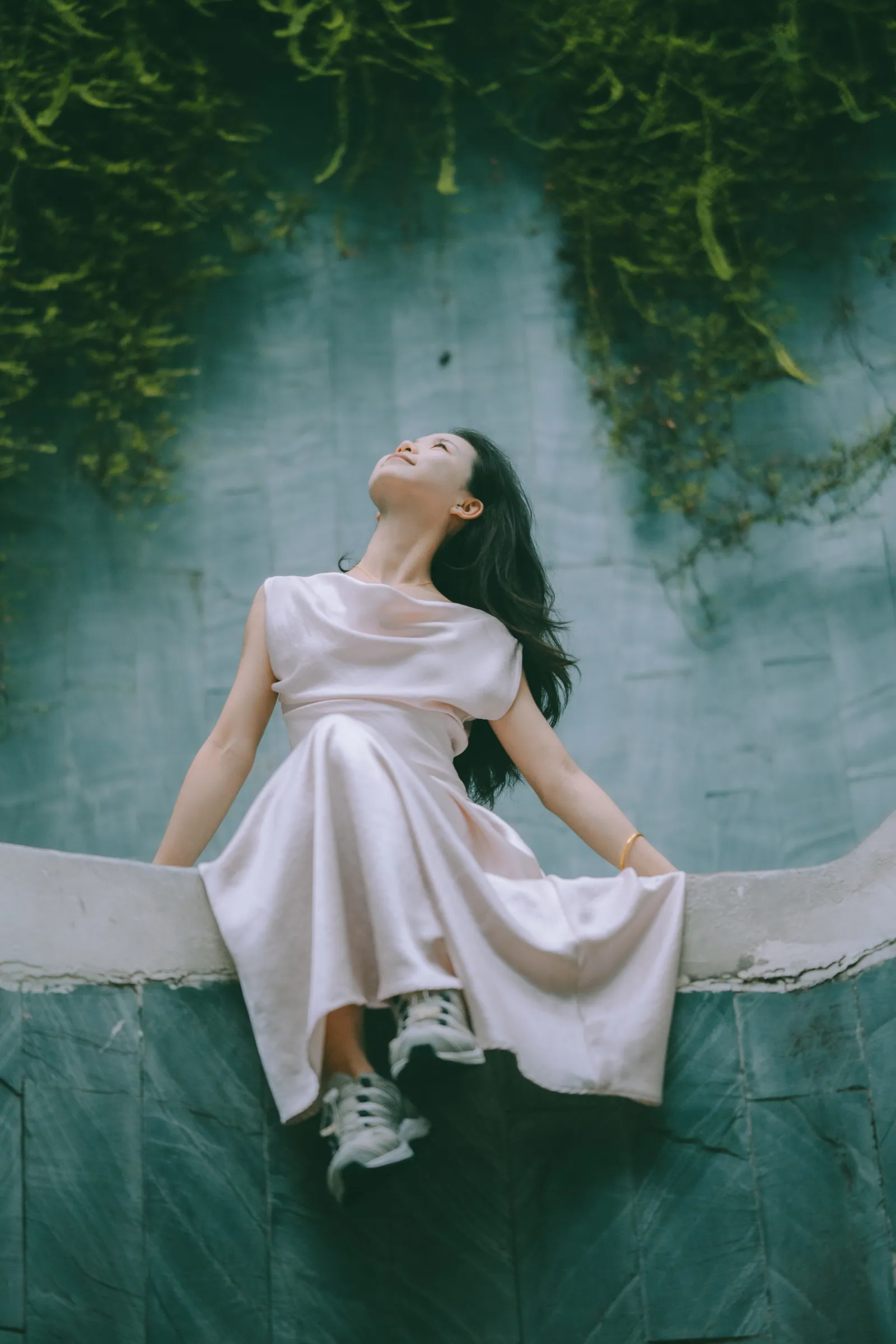 Woman in a pale pink satin dress sitting on a wall under hanging ferns, Fort Canning Park photoshoot, Singapore