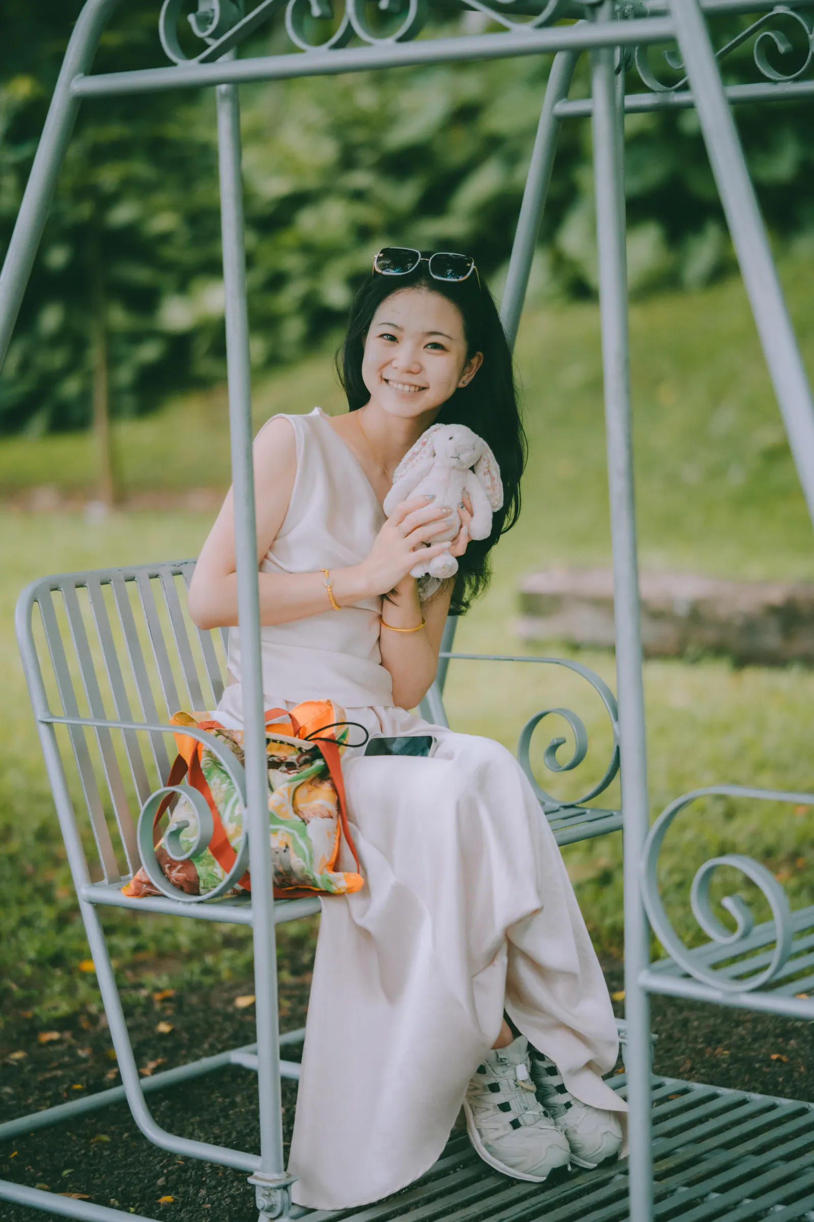 Woman in a cream dress on a garden swing holding a bunny plush, green moody portrait at Fort Canning Park