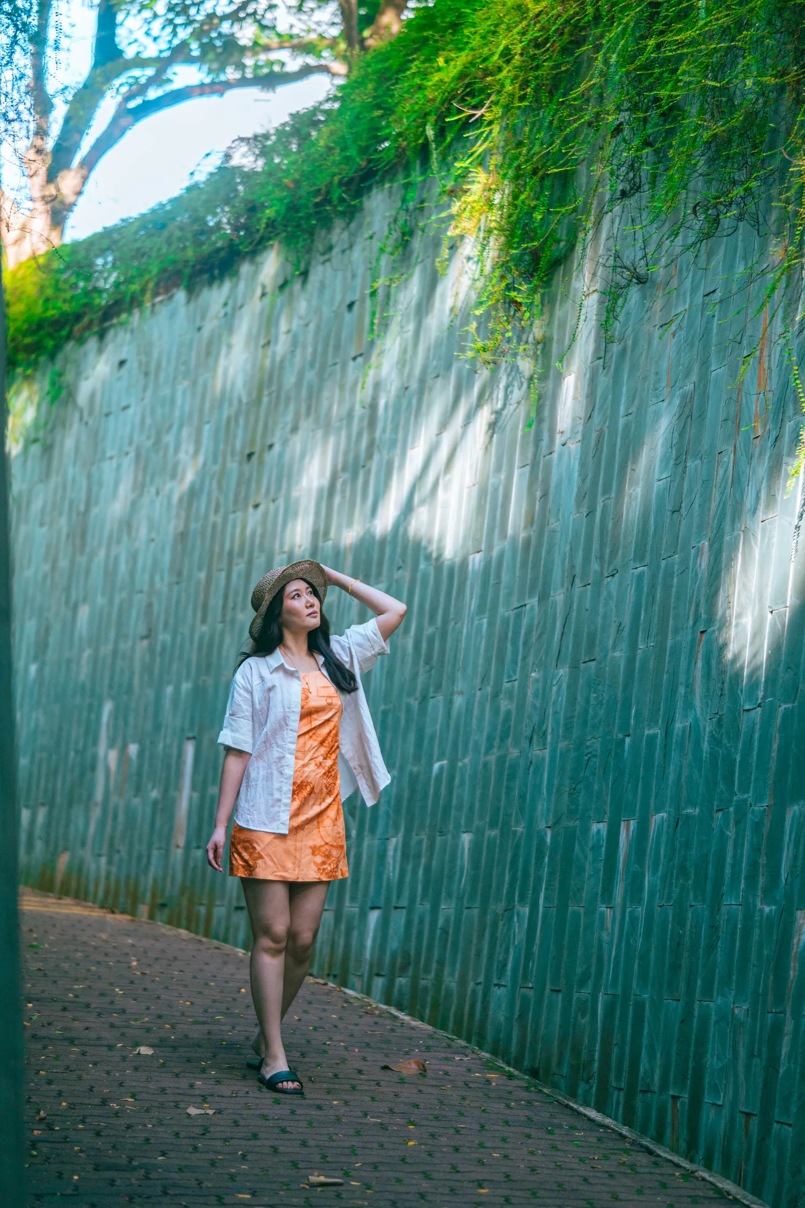 Woman in an orange dress and white shirt walking along a curved green tiled wall with plants hanging over it, Fort Canning Park