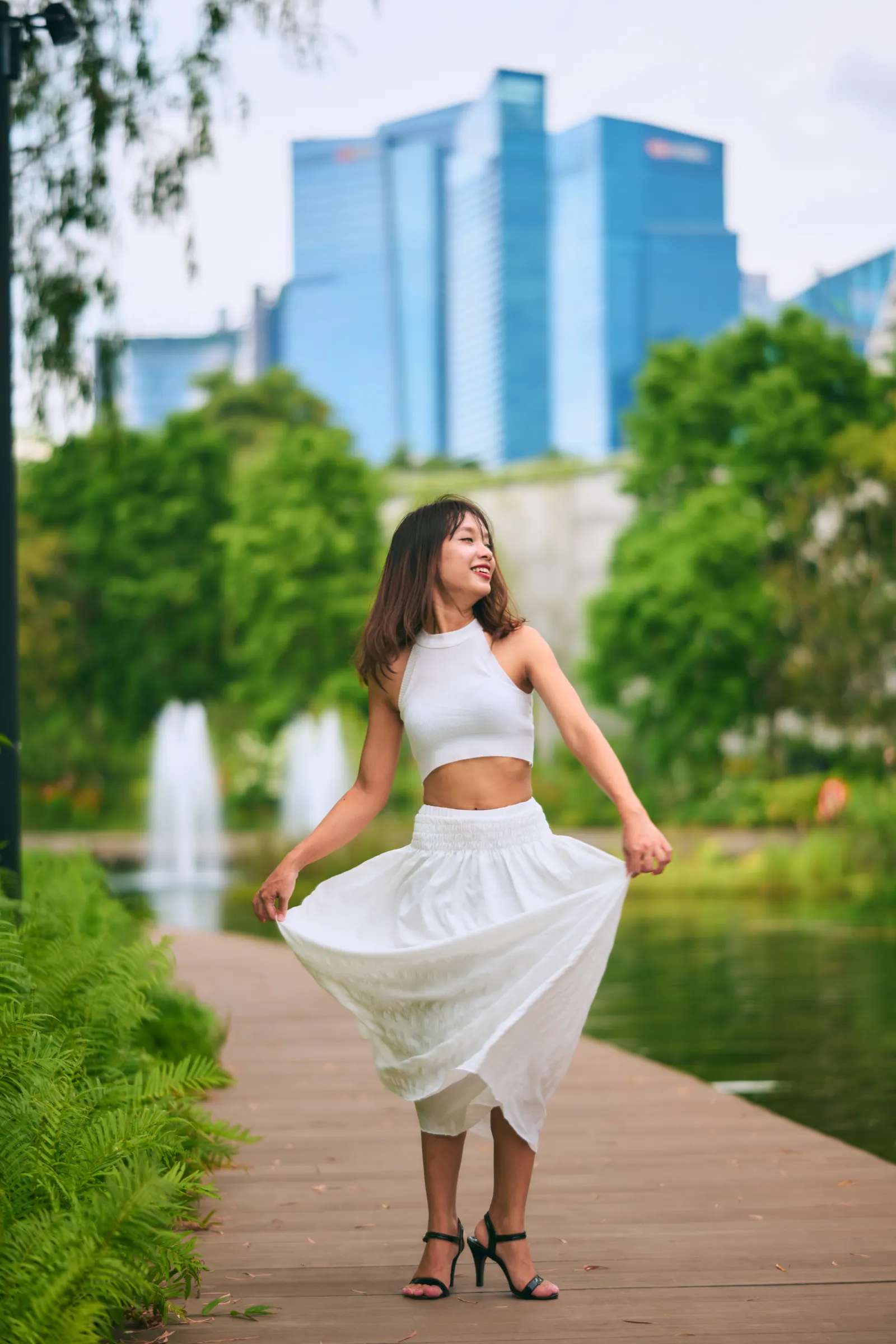 Woman twirling a long white skirt on a wooden boardwalk with fountains and the city skyline behind, Gardens by the Bay