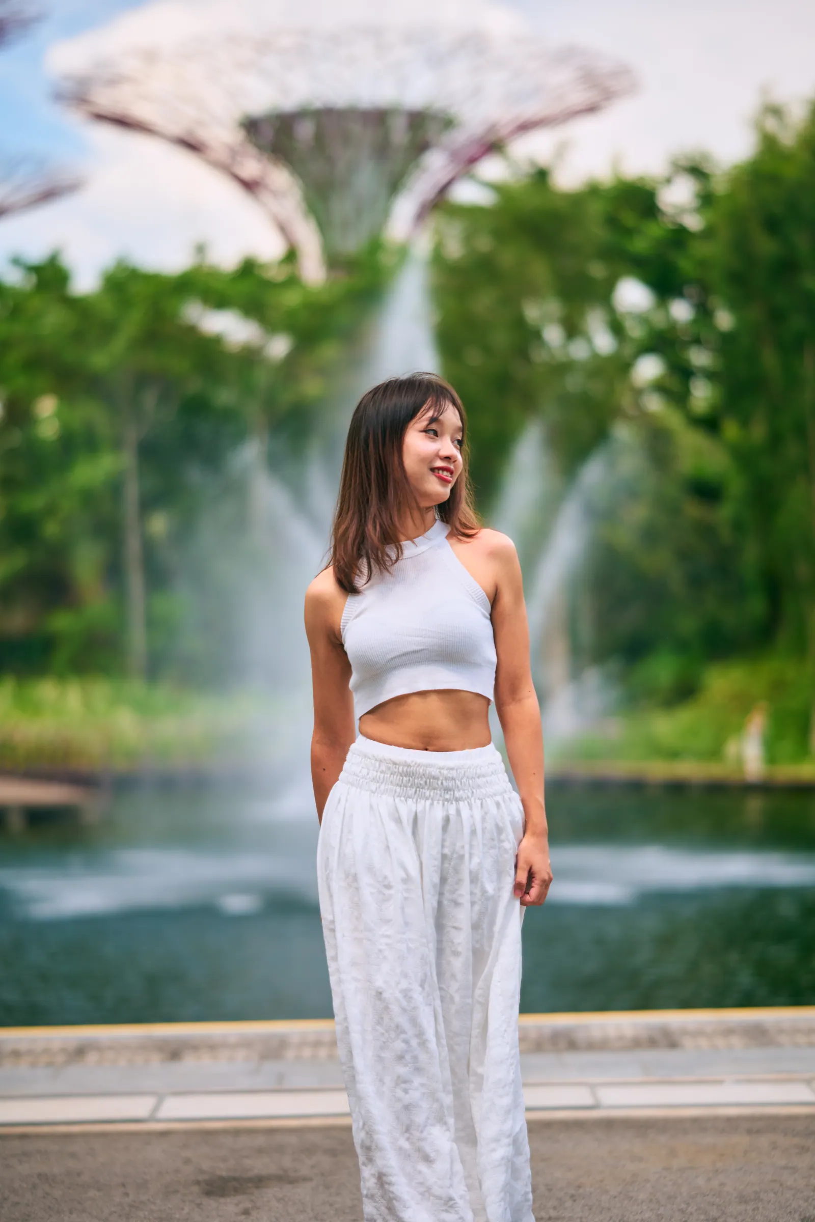 Woman in a white crop top and white trousers smiling beside a lake fountain, Gardens by the Bay photoshoot, Singapore