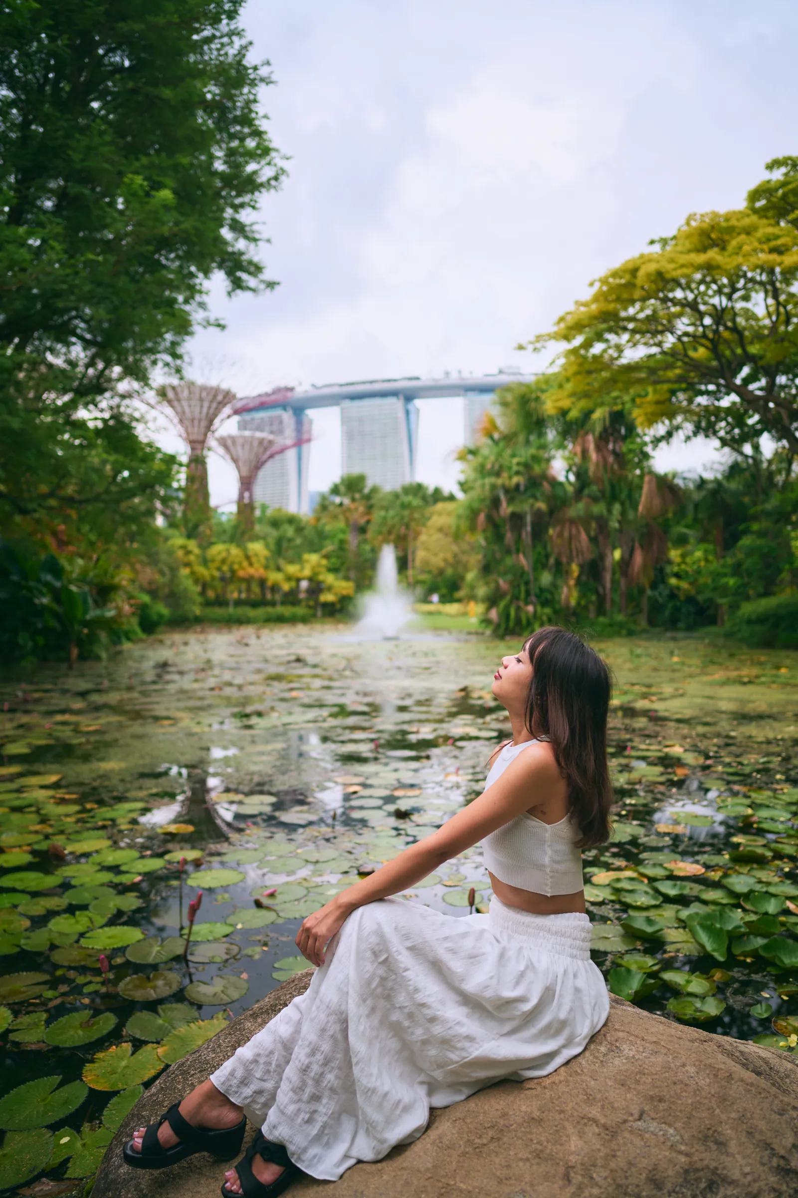 Woman in white sitting on a rock by a lily pond, with Supertrees and Marina Bay Sands in the distance, Gardens by the Bay