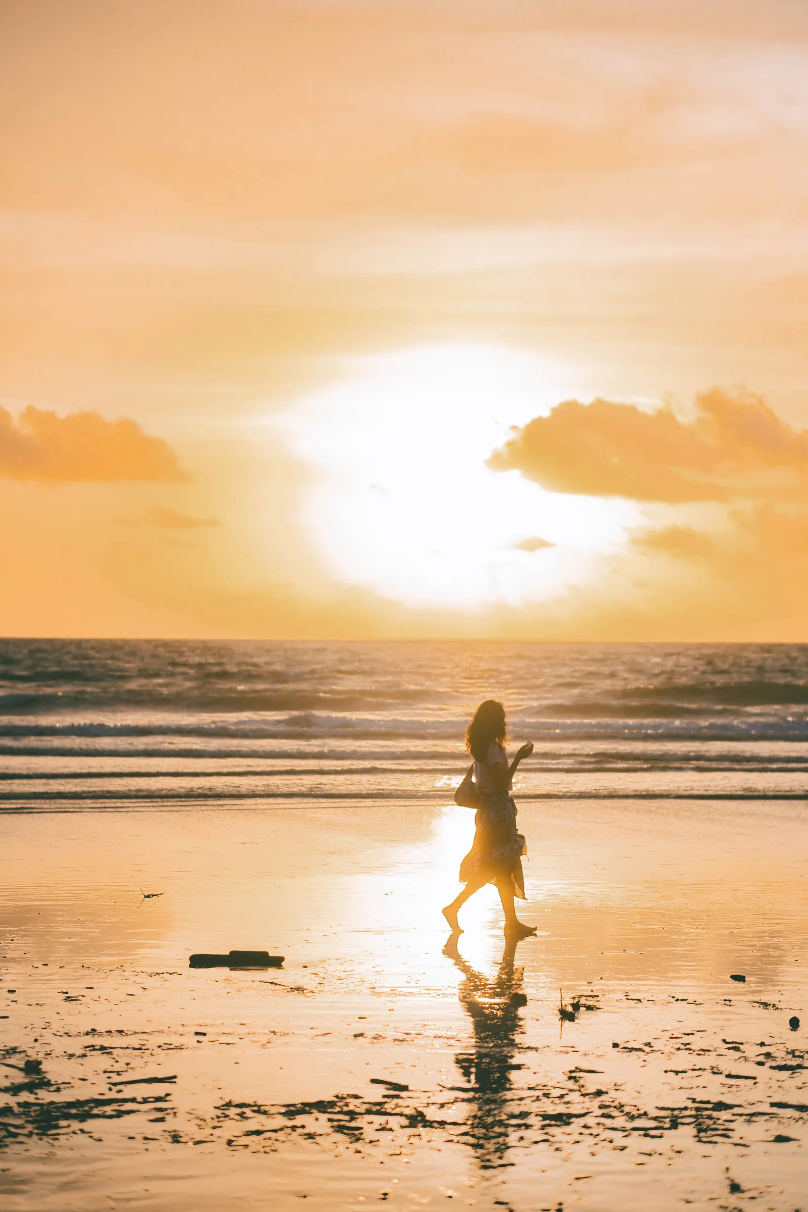Silhouette of a woman walking along a wet beach at sunset, the sun breaking through the clouds