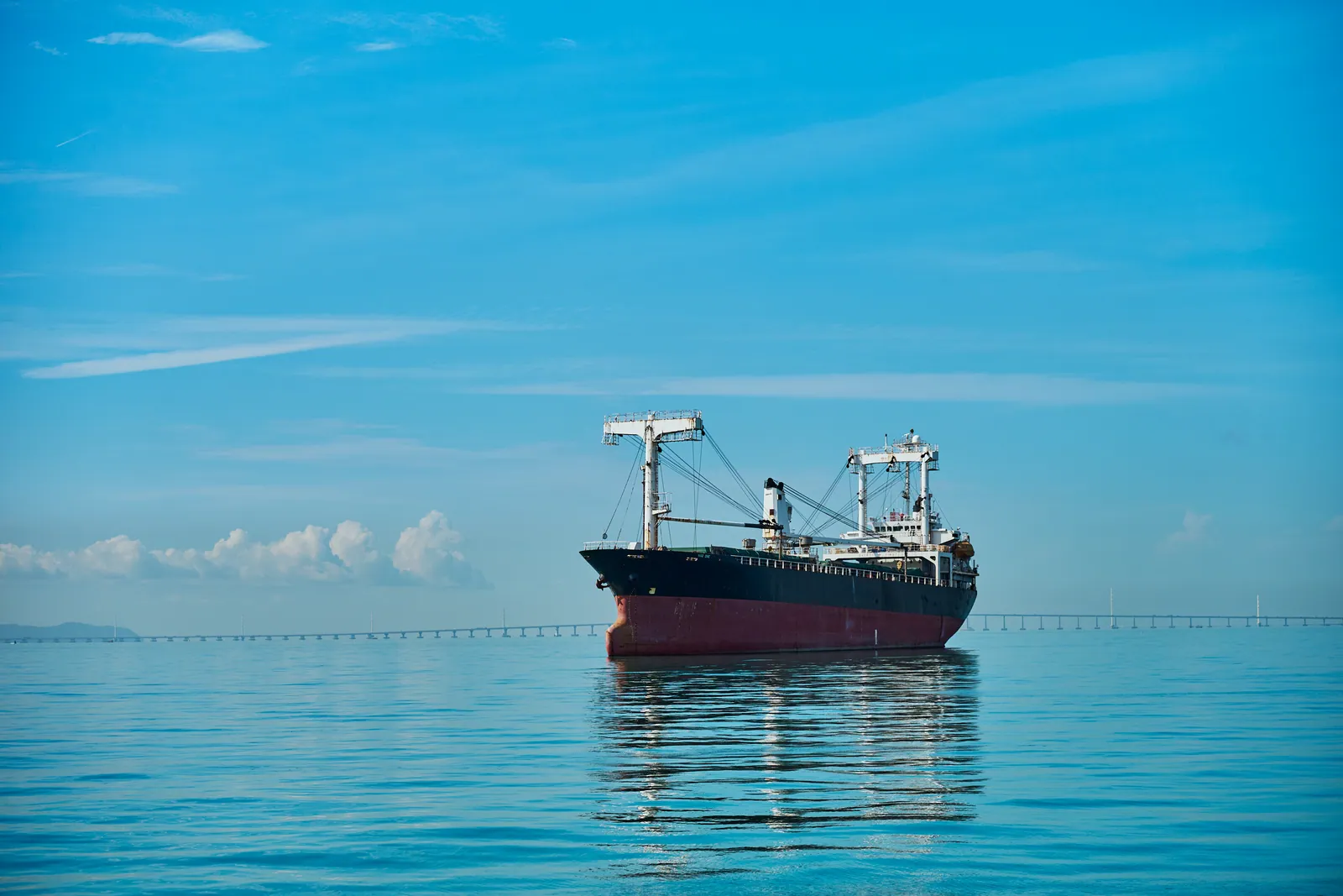 Cargo ship on a perfectly calm sea under a clear blue sky, with a long bridge on the horizon