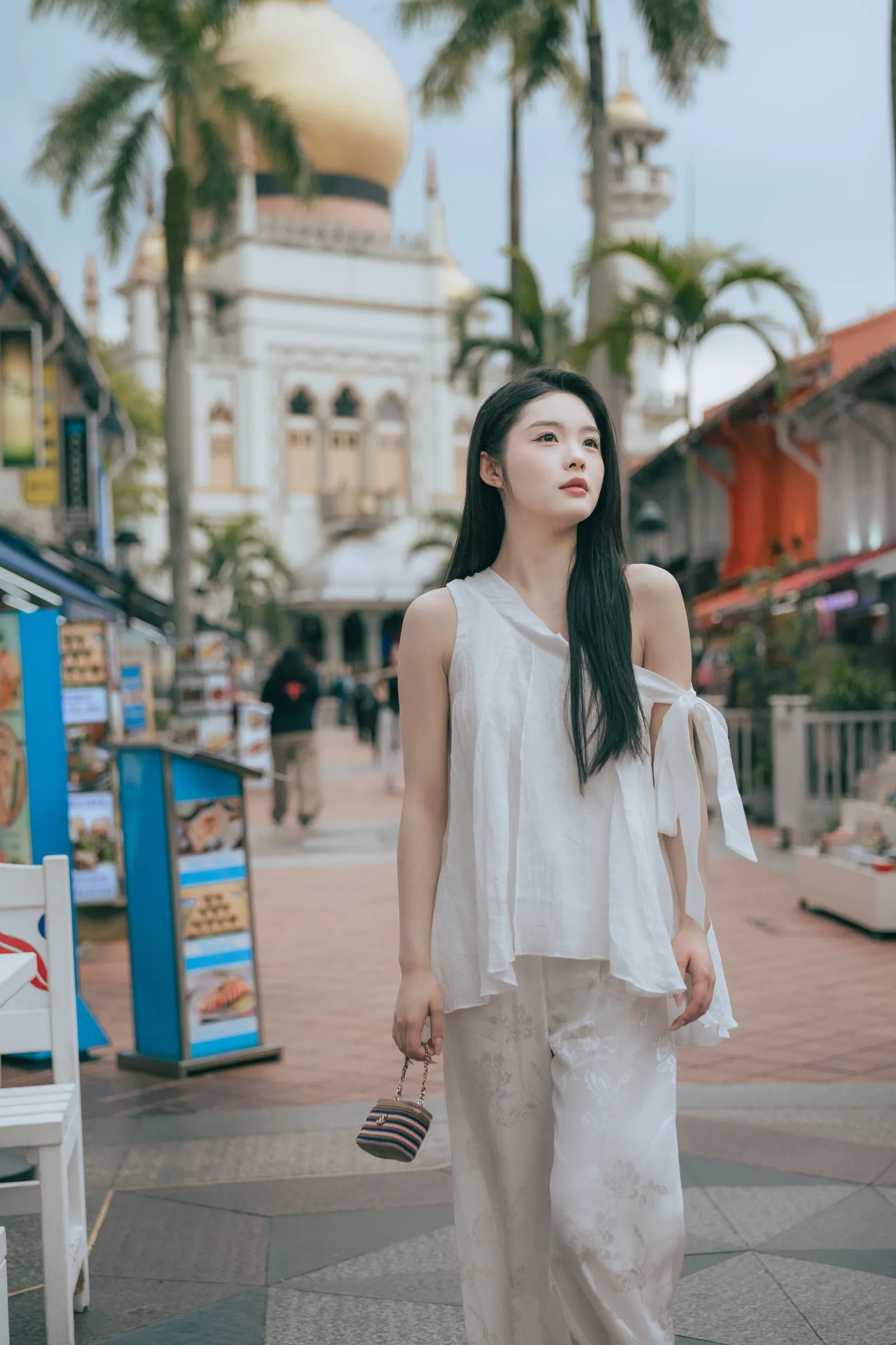 穿白色上衣的女生站在棕榈树成排的巴梭拉街上,身后是苏丹回教堂的金色圆顶,新加坡甘榜格南