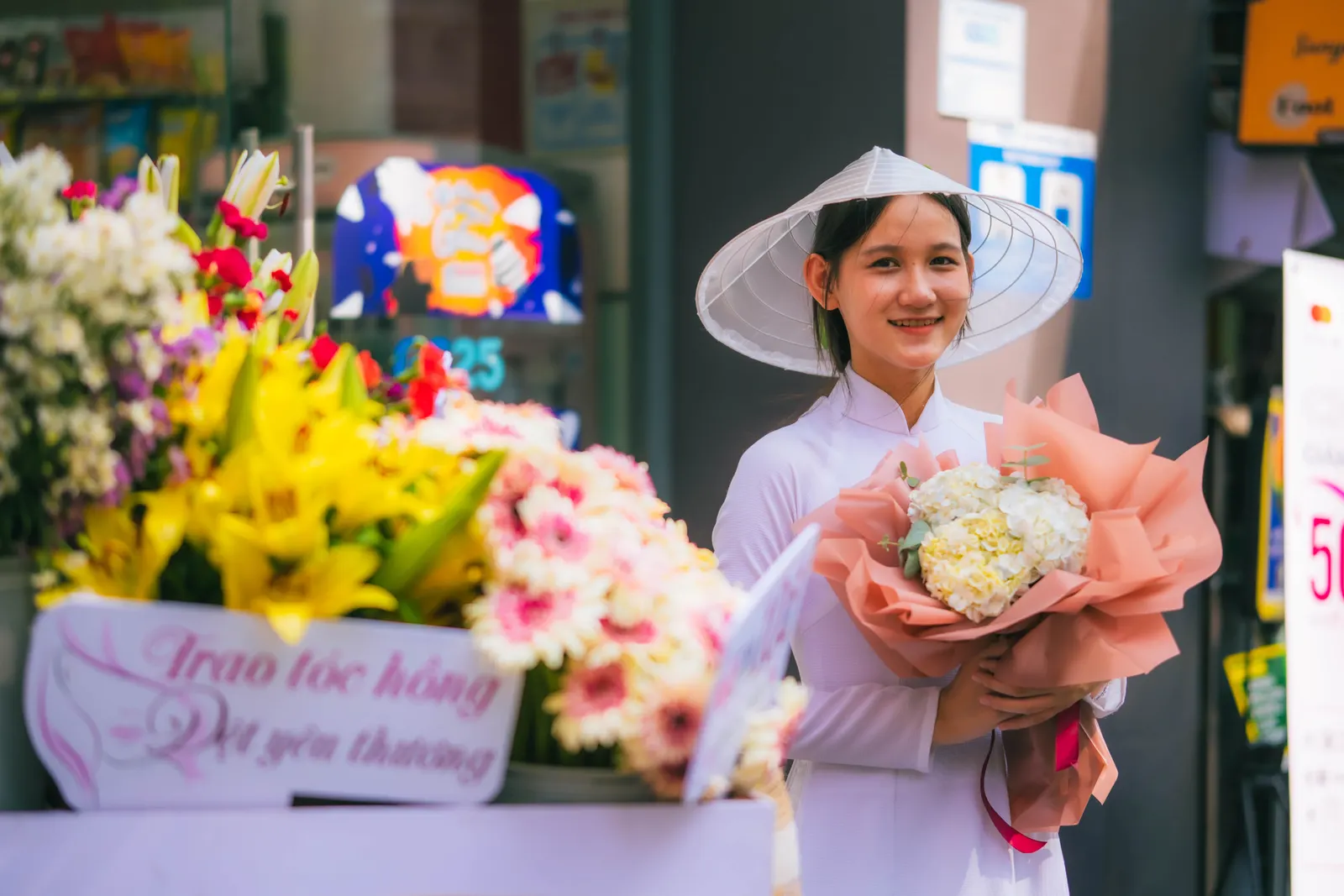 Woman in a white ao dai and conical hat holding a bouquet beside a street flower stall, Saigon