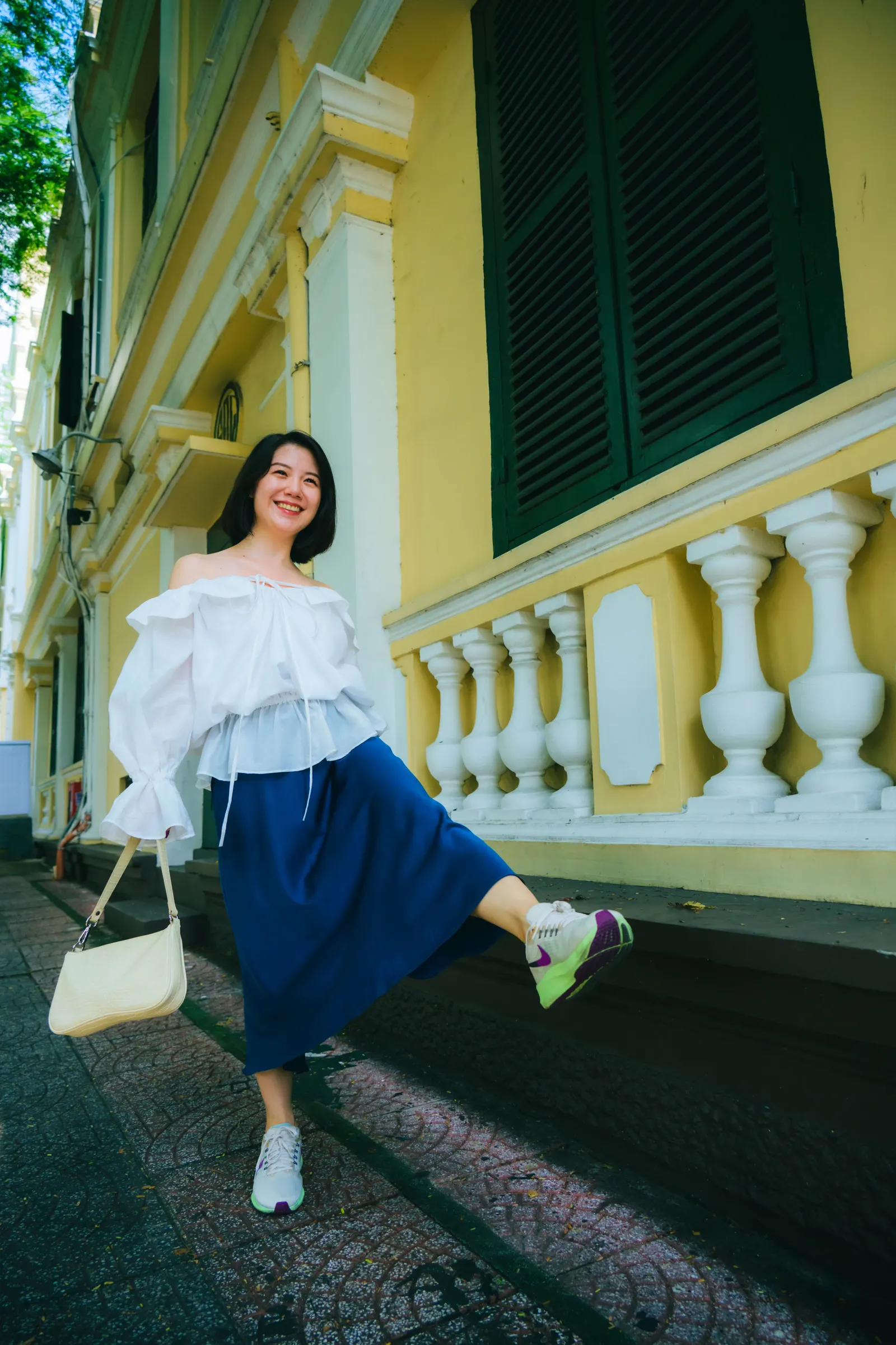 Woman laughing and kicking one leg up beside a yellow colonial wall with white balusters, Saigon
