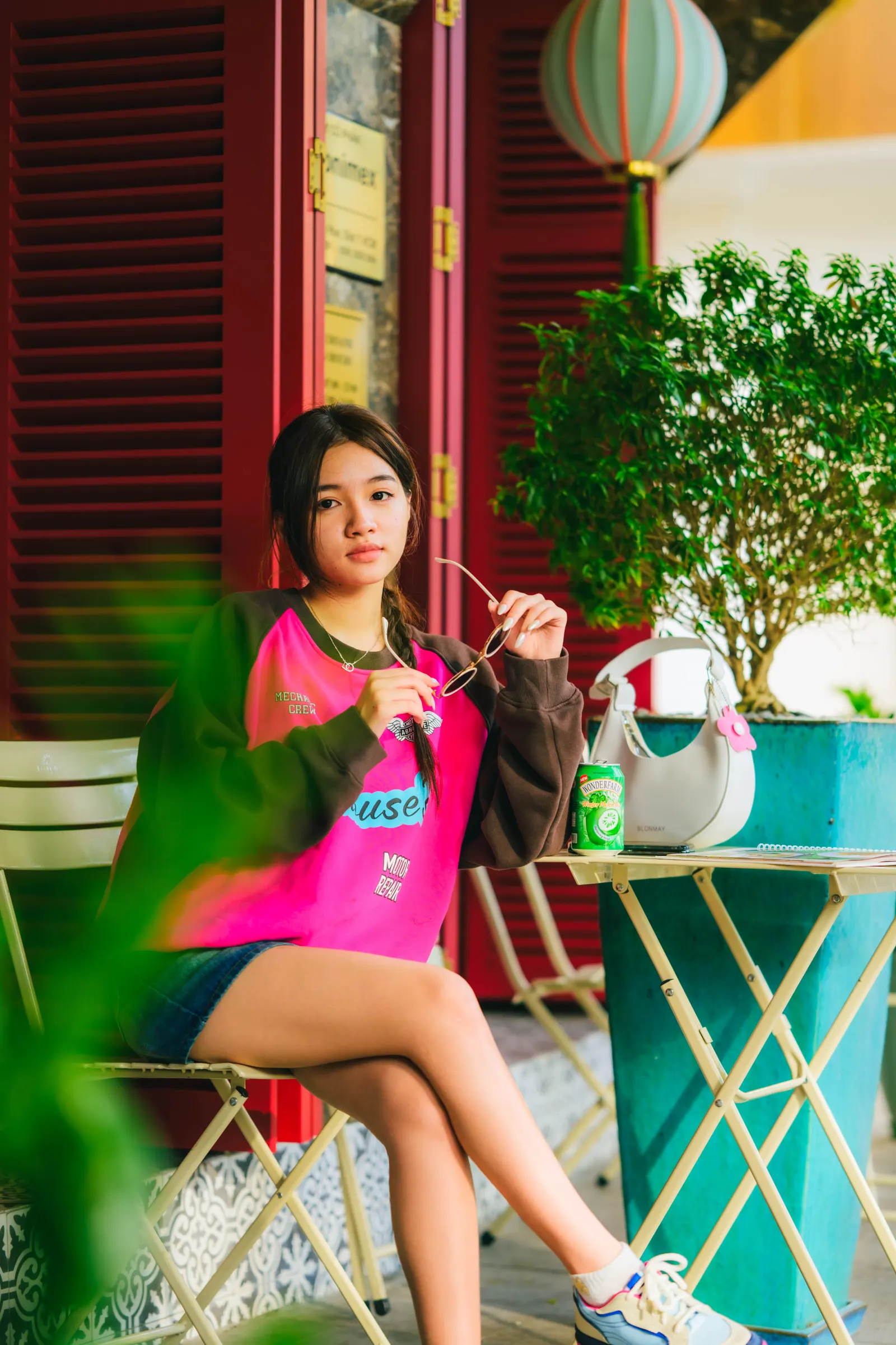 Young woman in a pink sweatshirt sitting at a café table by red shutters and a lantern, Ho Chi Minh City