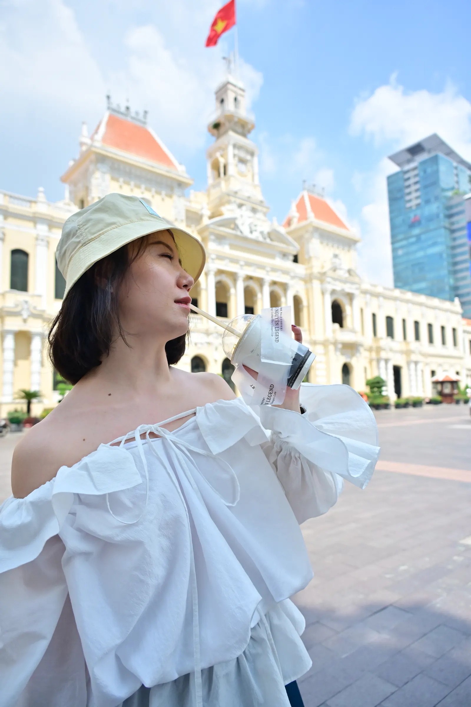 Woman sipping an iced coffee in front of Ho Chi Minh City Hall under a blue sky