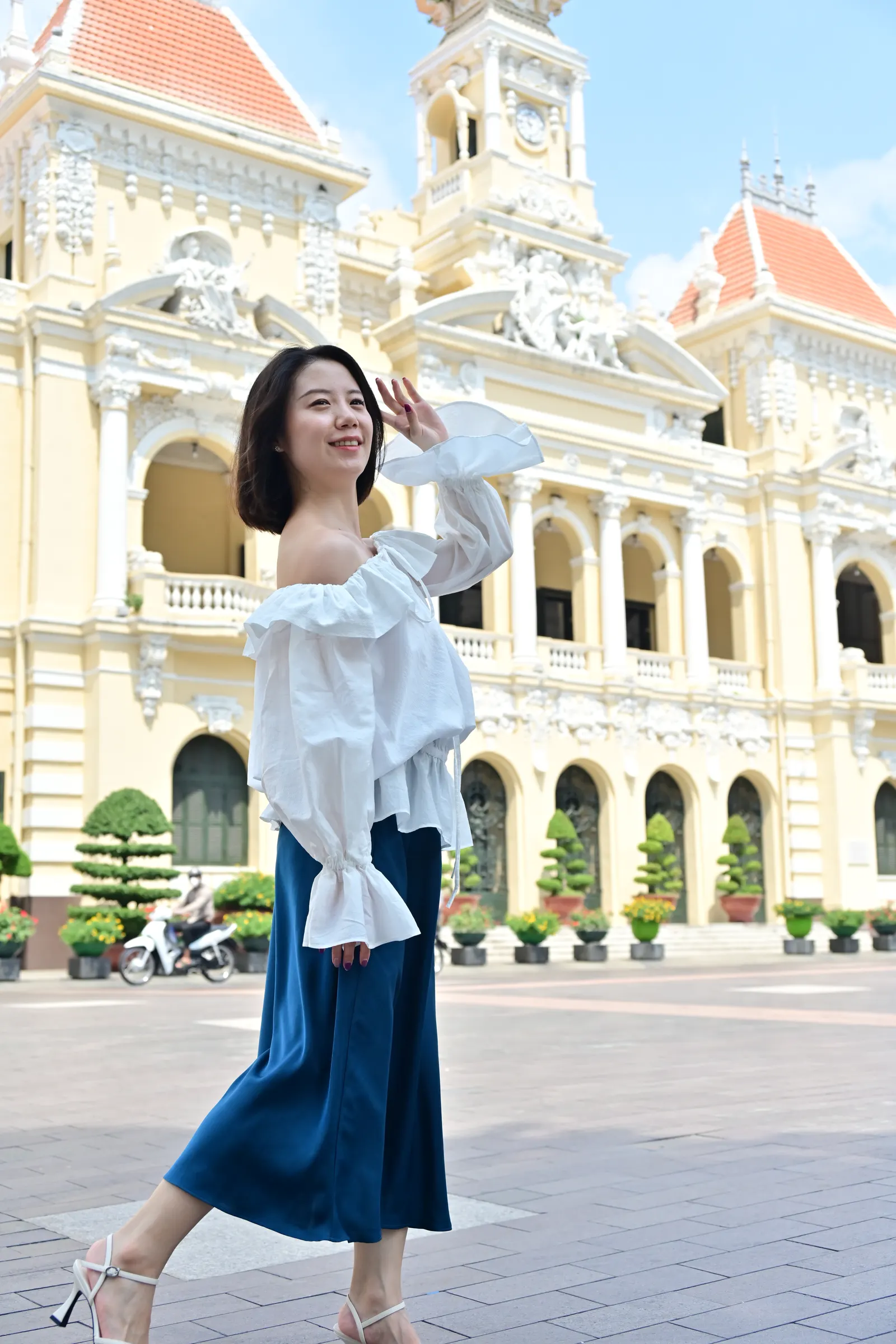Woman in a white ruffled top and blue skirt walking in front of the yellow colonial City Hall in Ho Chi Minh City