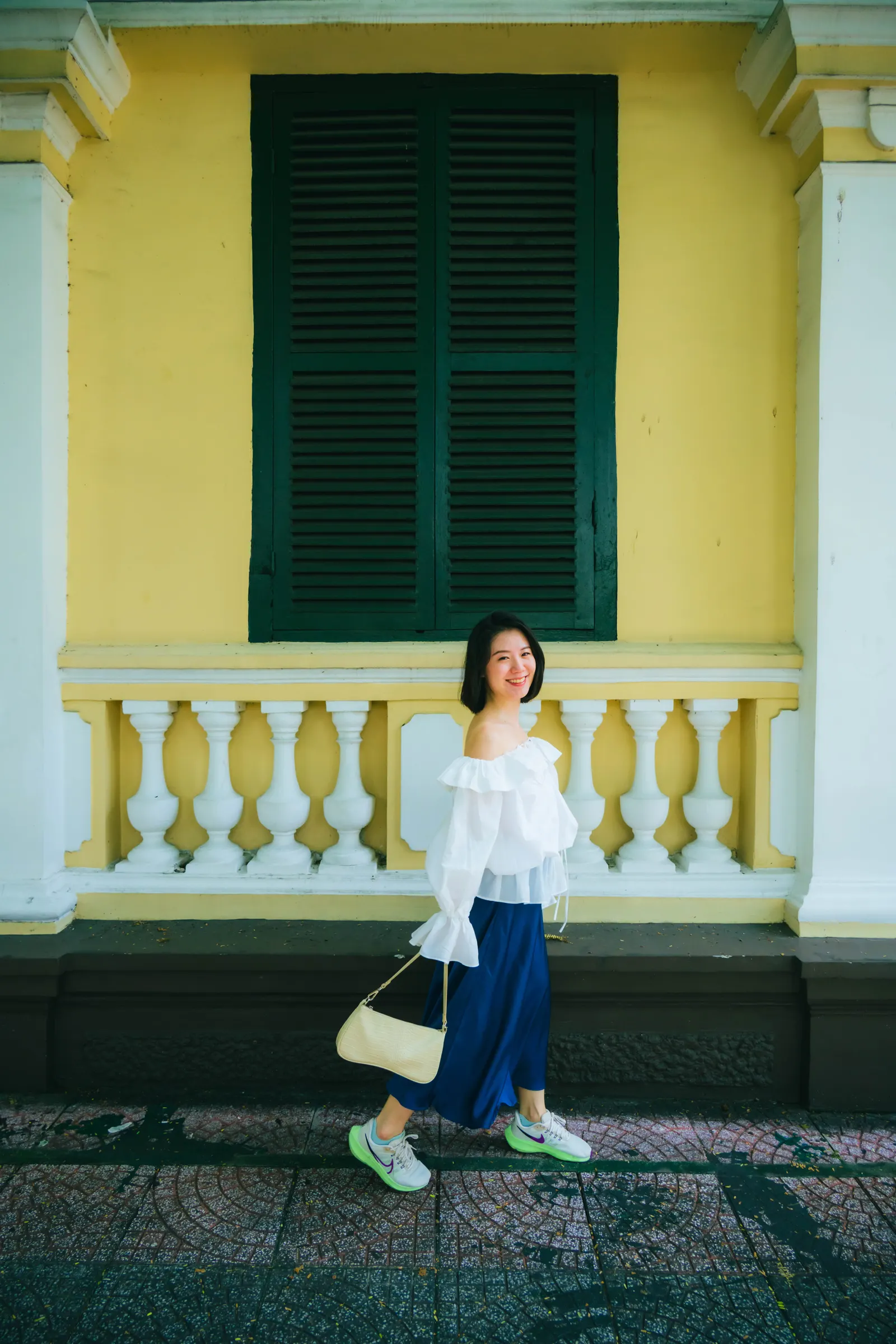Woman smiling as she walks past a yellow French colonial building with green shutters, Saigon