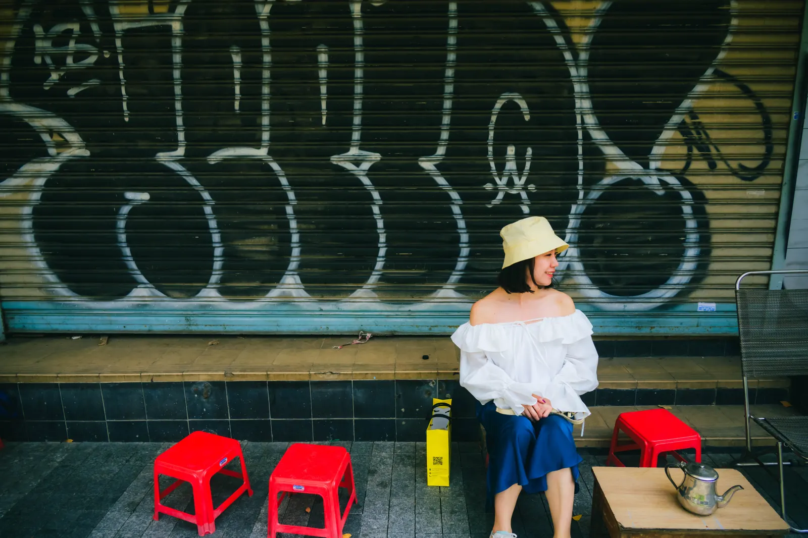 Woman in a white off-shoulder top and bucket hat sitting by red plastic stools in front of a graffiti shutter, Ho Chi Minh City