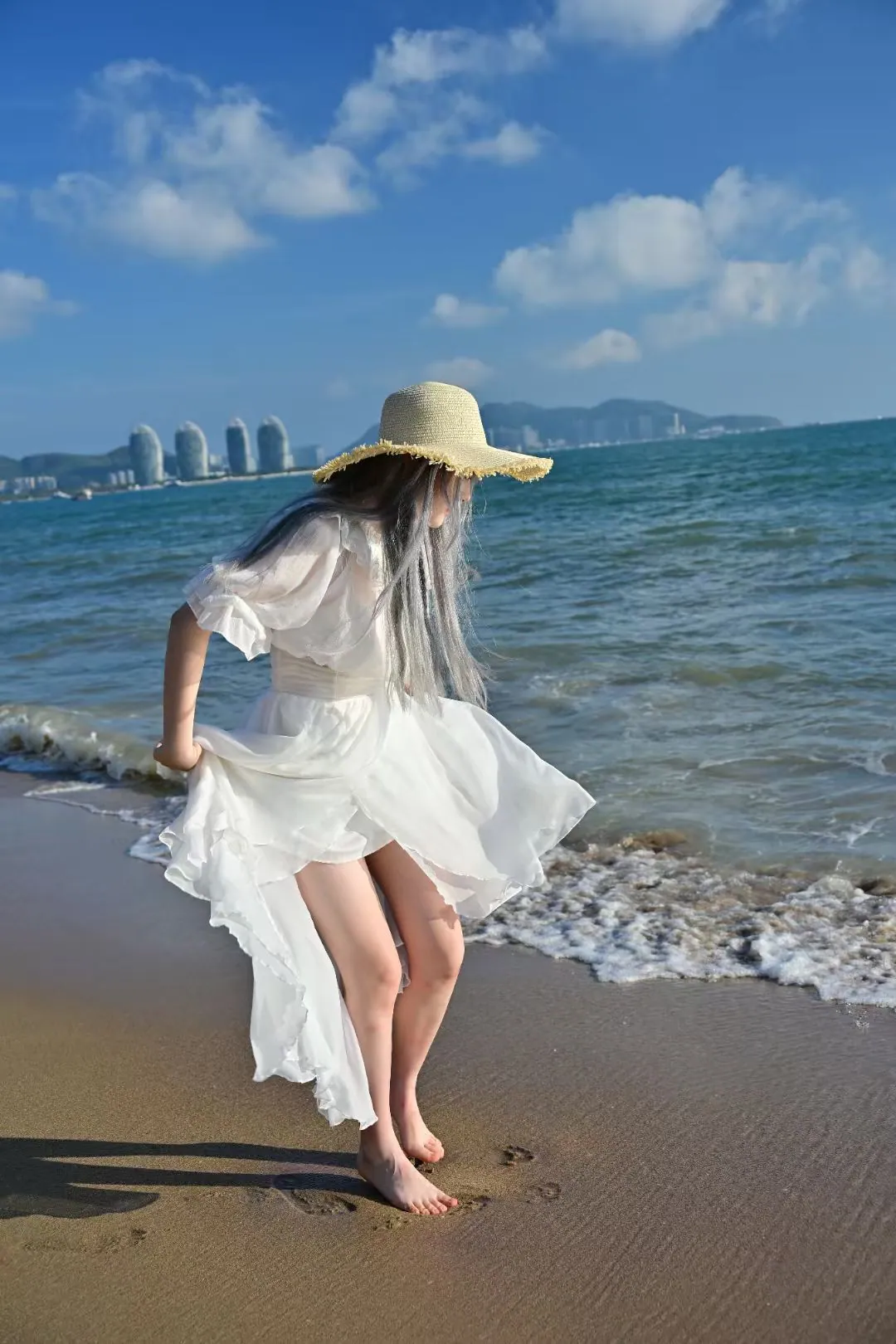 Woman in a white dress and straw hat stepping barefoot on wet sand as a wave comes in, Sanya beach photoshoot