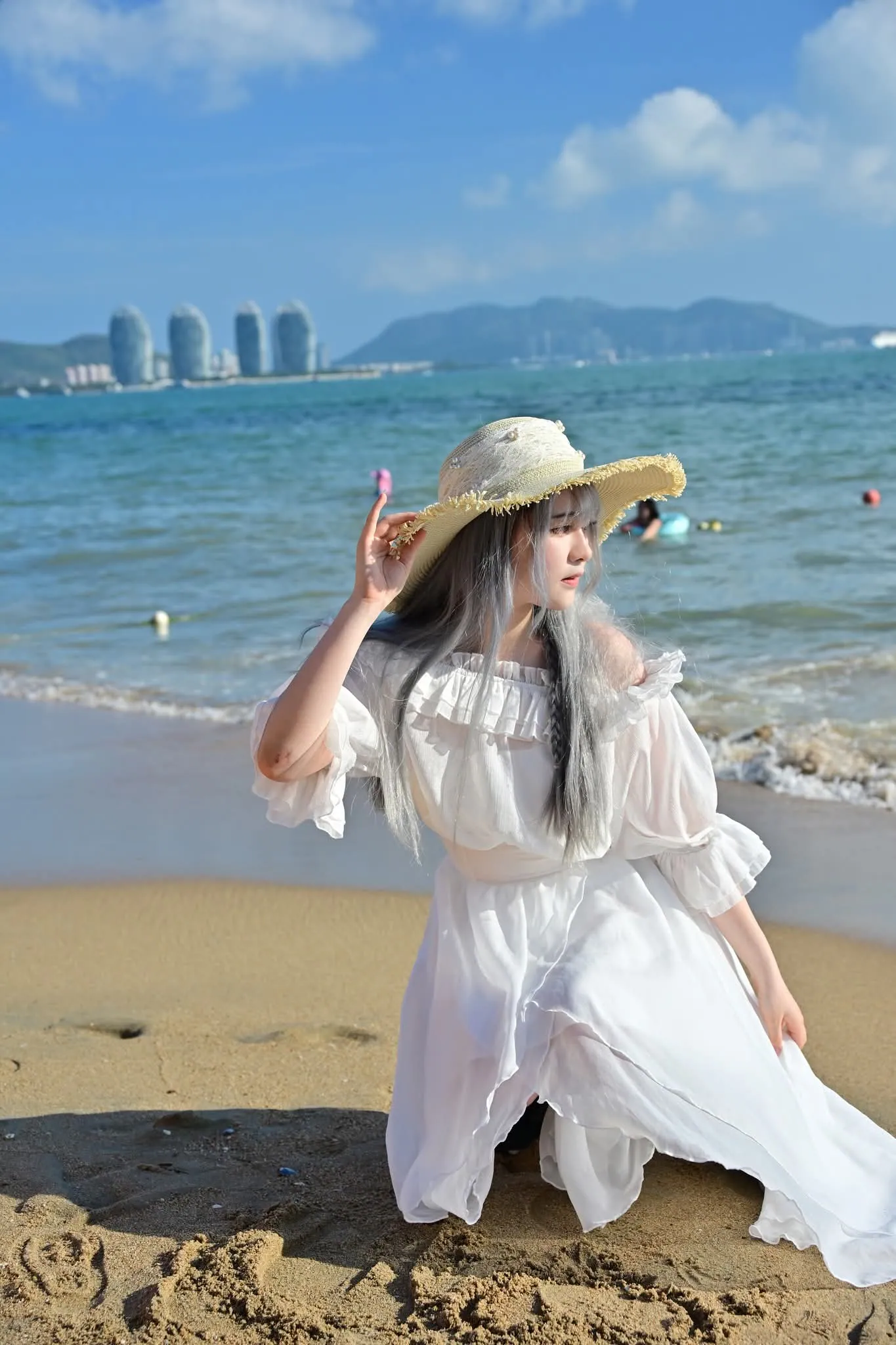 Woman in a white dress kneeling on the sand and holding her straw hat, with the sea and city towers behind her in Sanya
