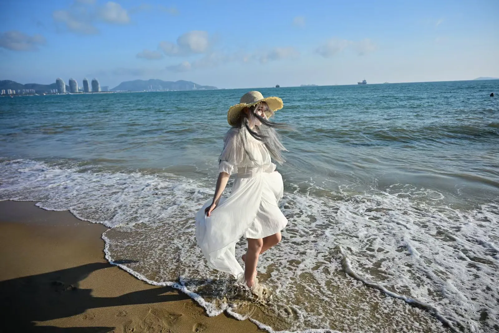 Woman in a white dress and straw hat walking through the waves and looking back, beach photoshoot in Sanya, China