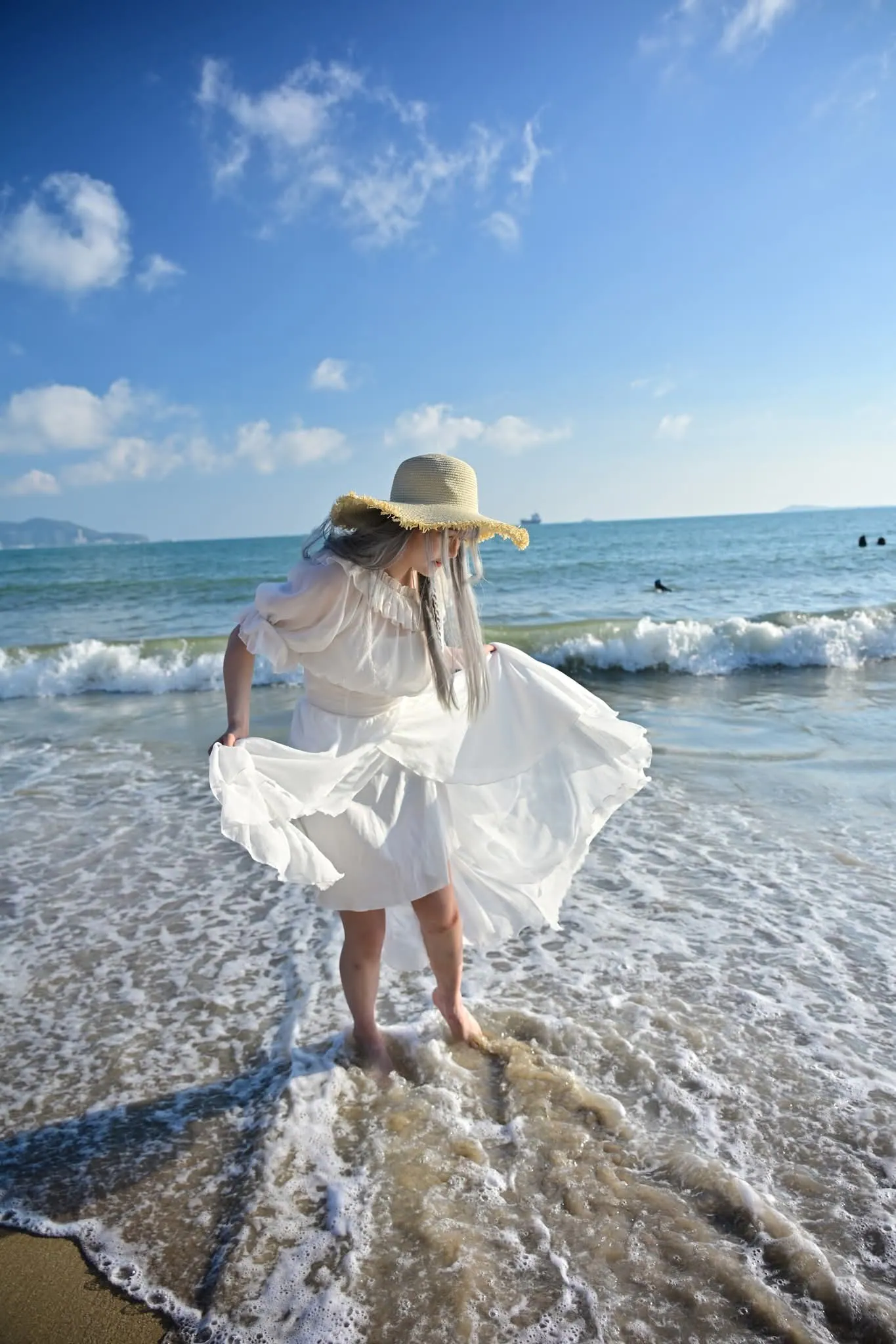 Woman in a white dress and straw hat lifting her skirt as a wave washes over her feet, Sanya beach