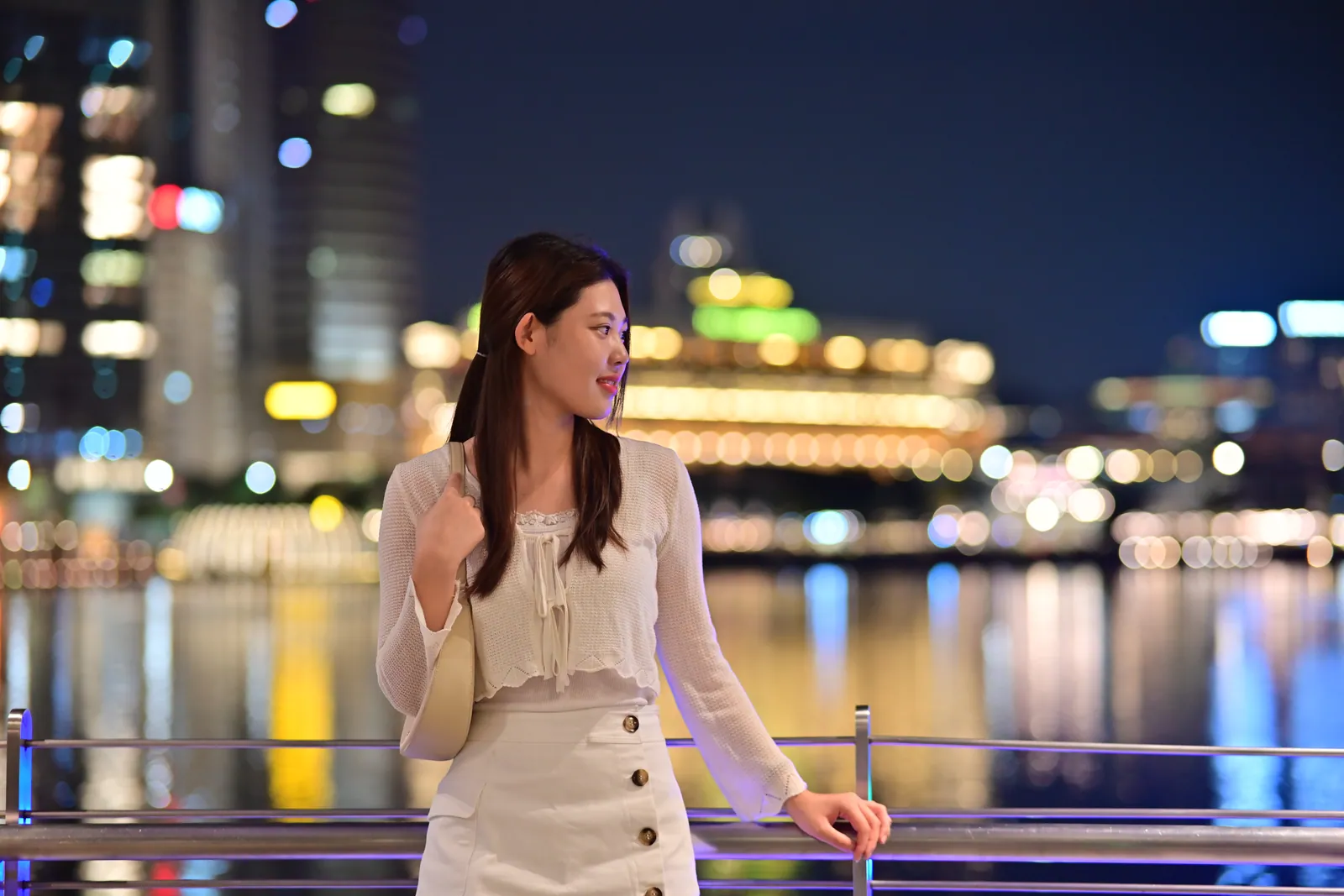 Woman in a cream cardigan and skirt leaning on a railing at night, with Marina Bay city lights softly blurred into bokeh