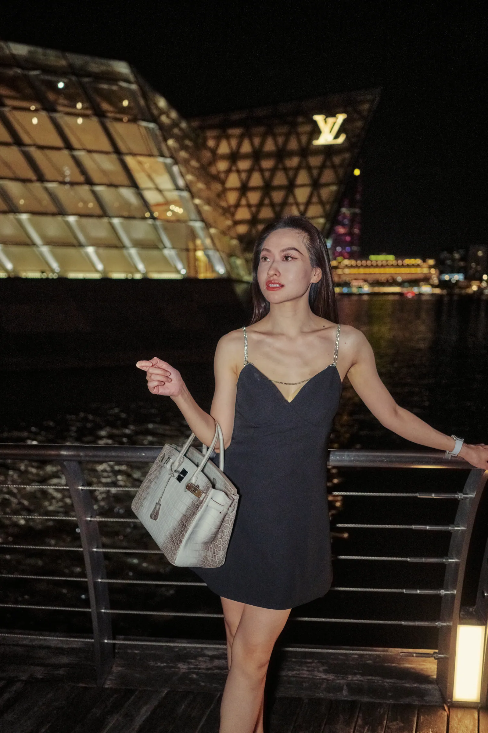 Woman in a black dress holding a handbag in front of the lit crystal pavilion on the water at Marina Bay at night