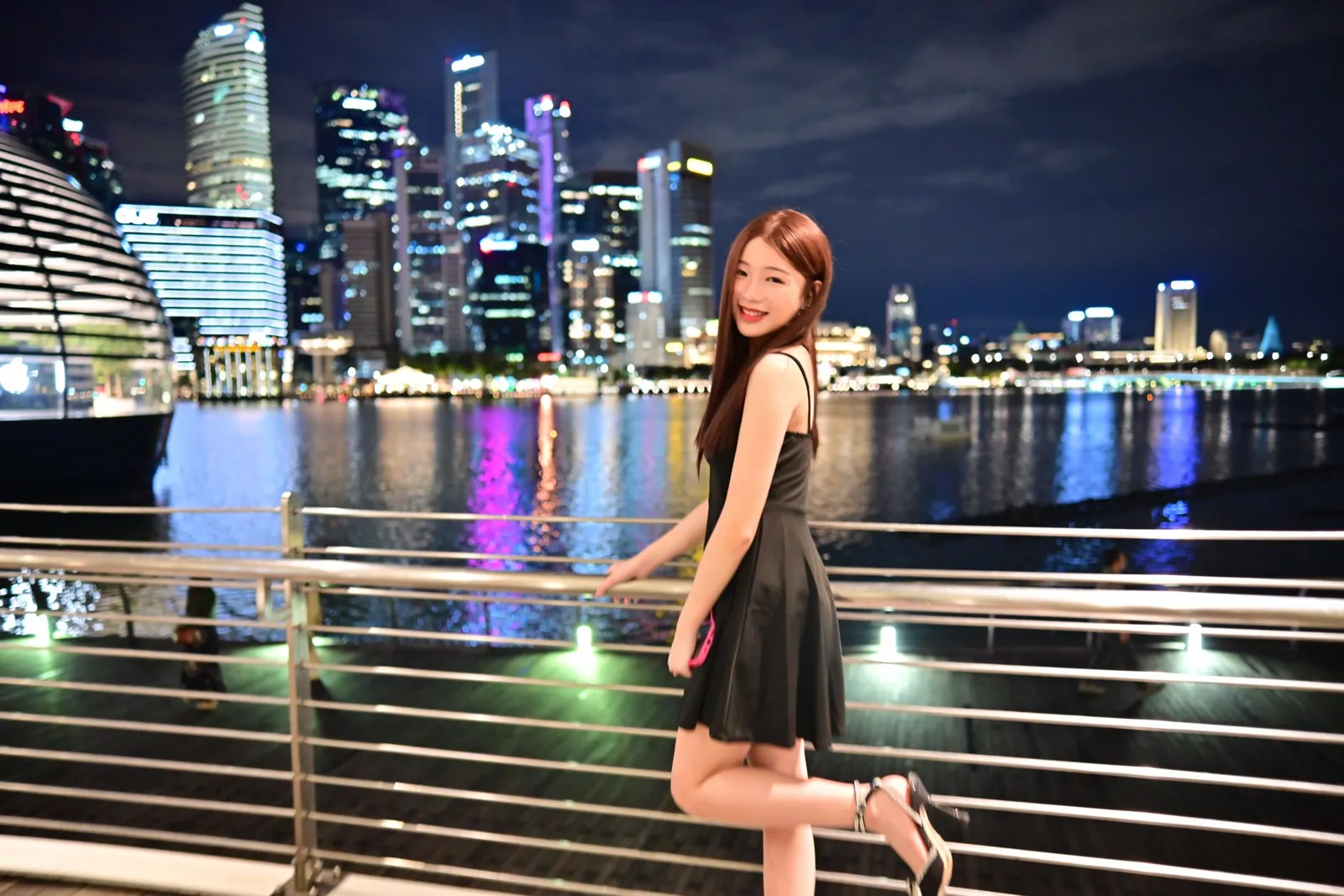 新加坡滨海湾夜景人像:穿黑色连衣裙的女生靠在栏杆边,身后是倒映在水中的城市天际线