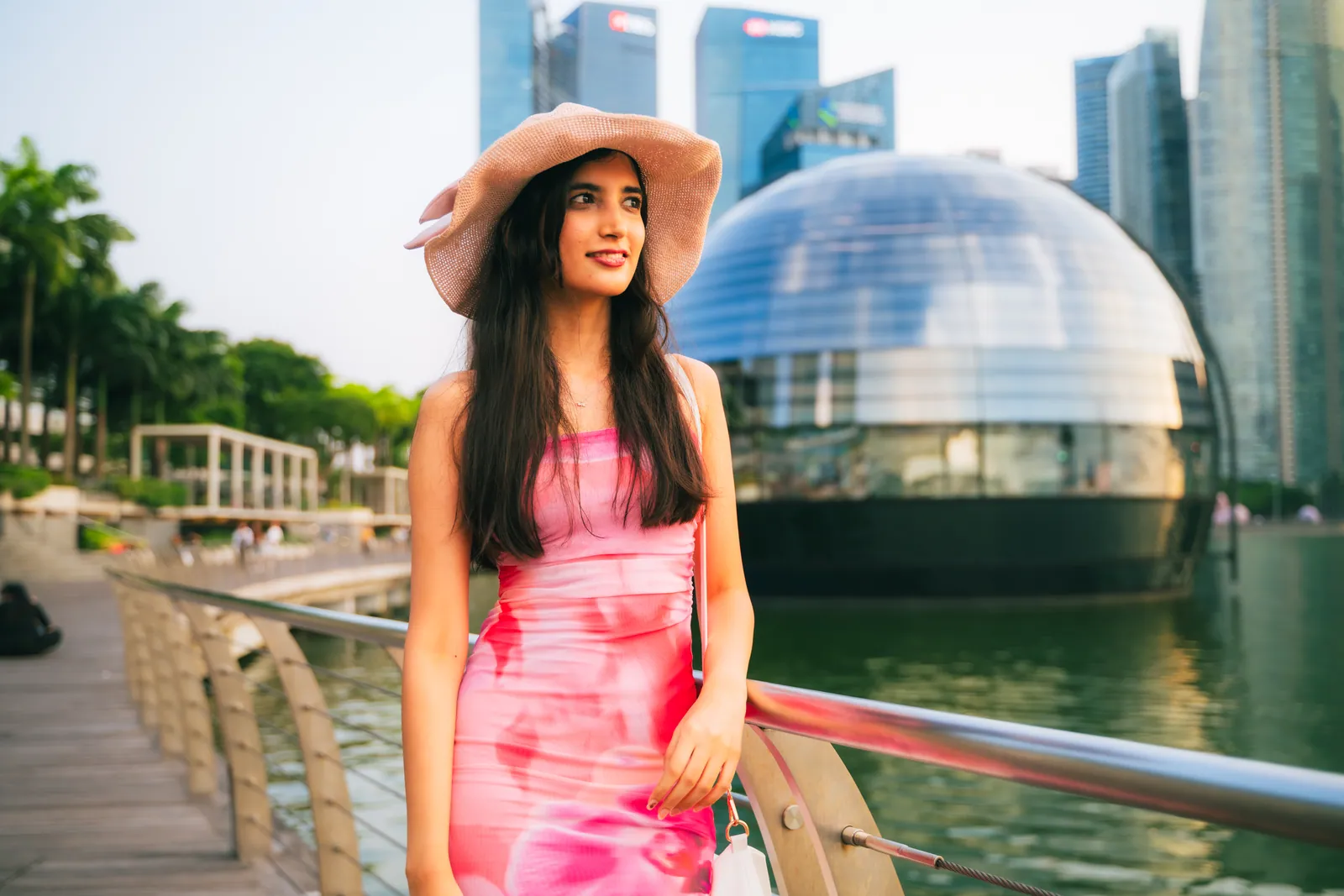 Woman in a pink dress and wide-brimmed hat by the Marina Bay promenade, with the glass Apple sphere and city towers behind