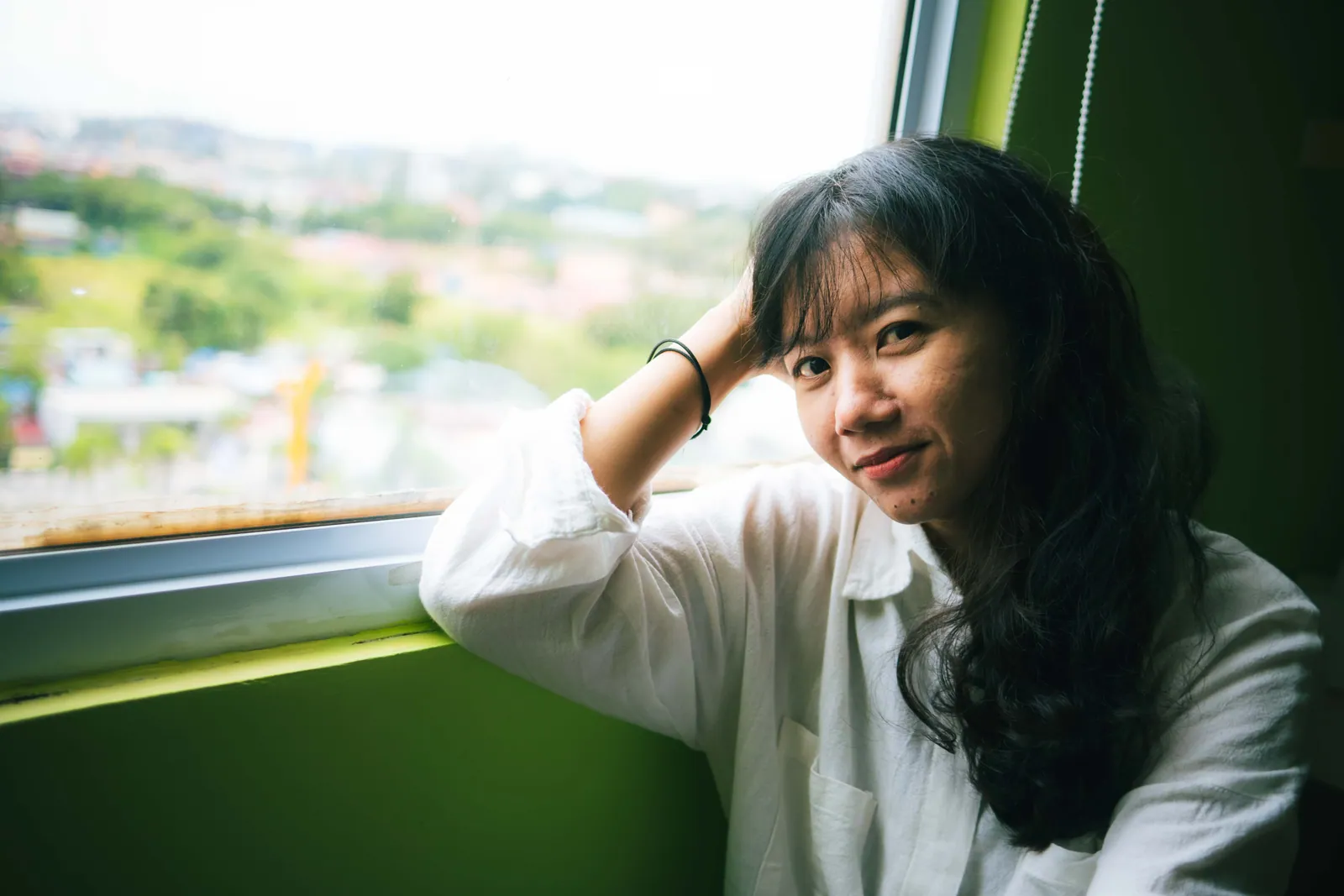 Woman in a white shirt resting her head on her hand by a window, looking at the camera with a soft smile