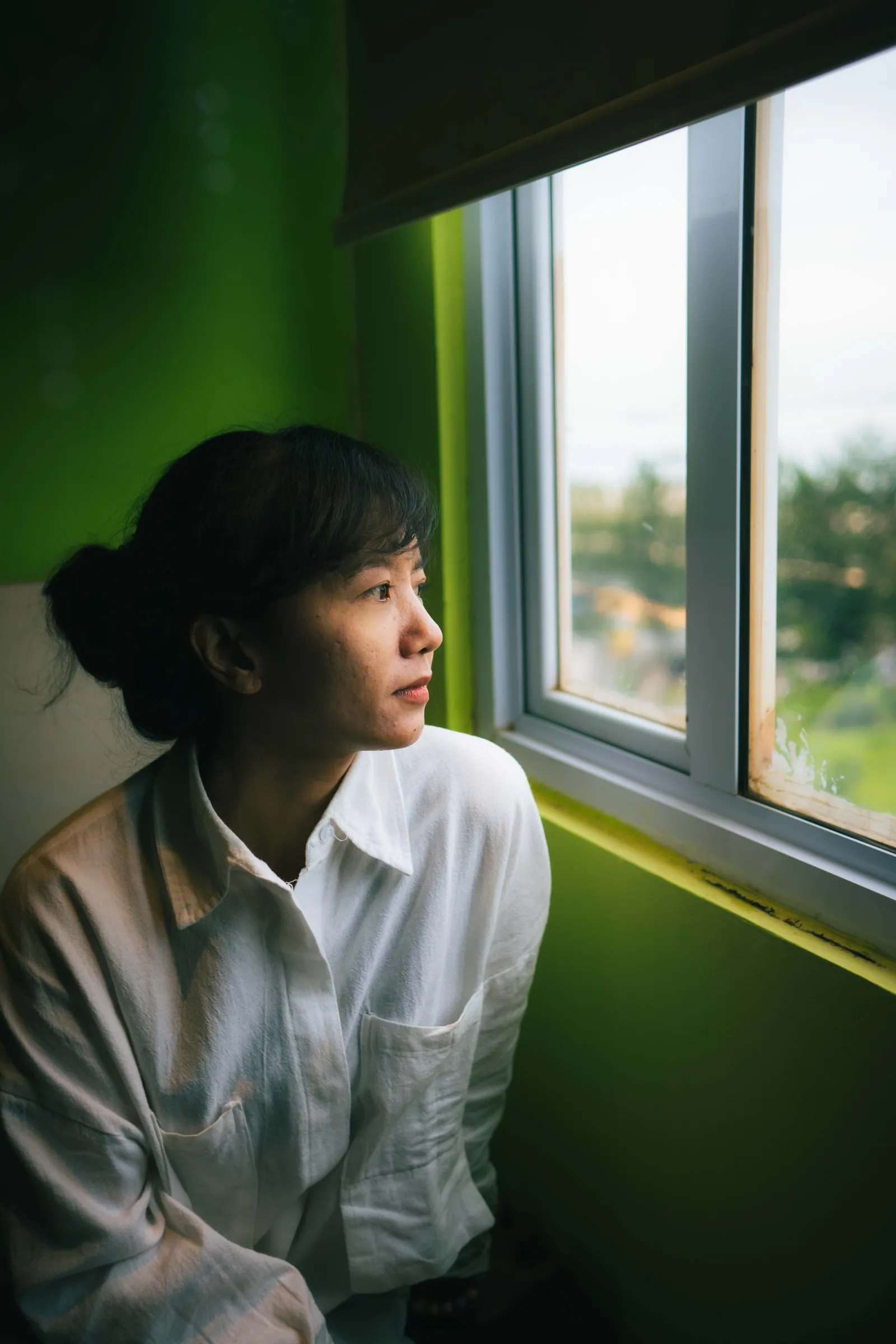 Woman in a white shirt gazing out of a window, soft light on her face against a green wall