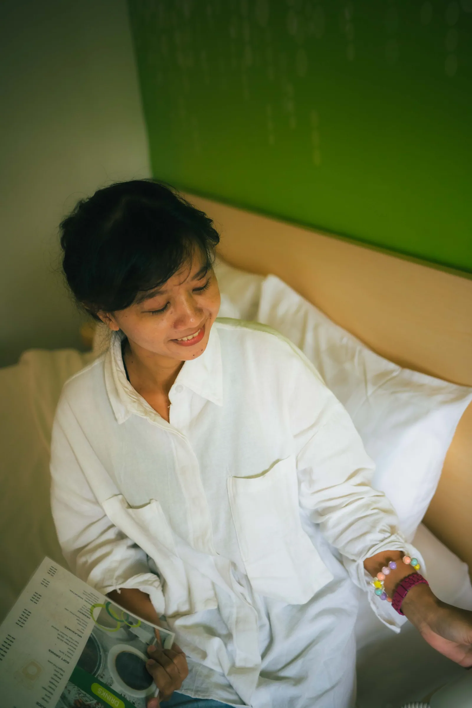 Woman in a white shirt sitting on a bed and smiling down, holding a menu, in a room with a green wall