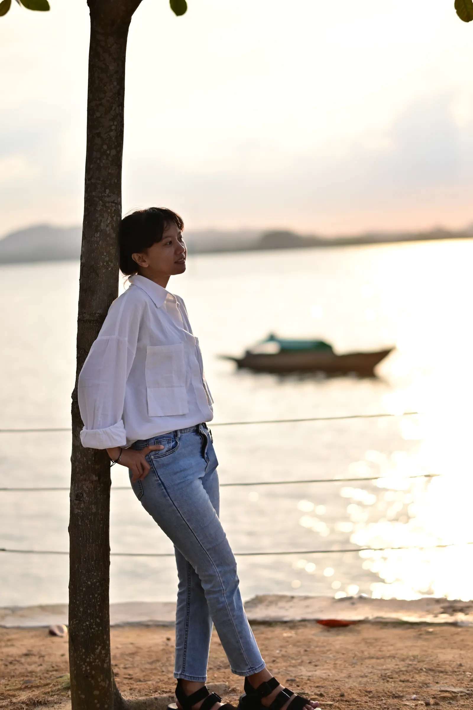 Woman in a white shirt and jeans leaning against a tree by the water at sunset, a boat on the sea behind her