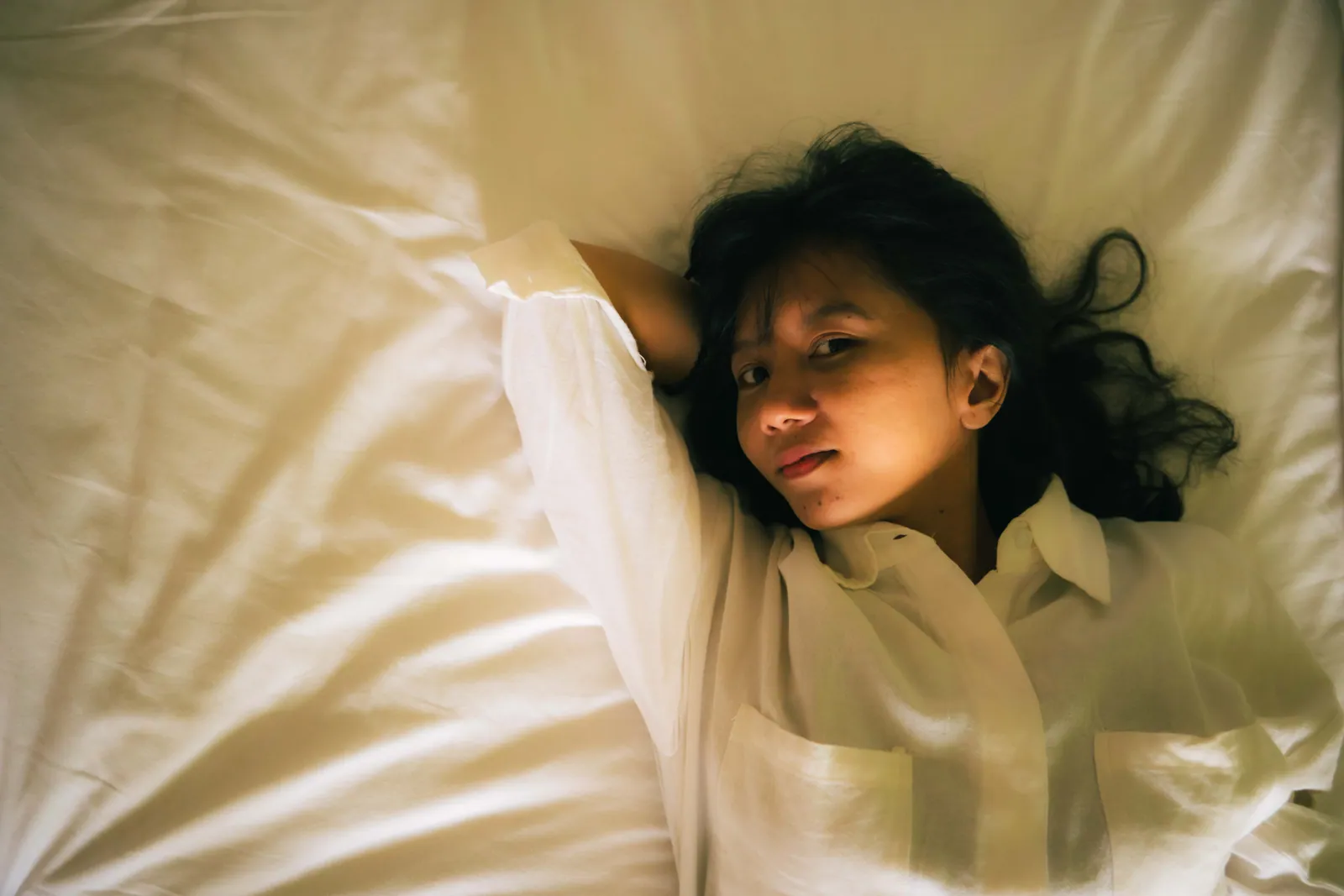 Woman in a white shirt lying on white sheets, looking up at the camera in warm, soft light