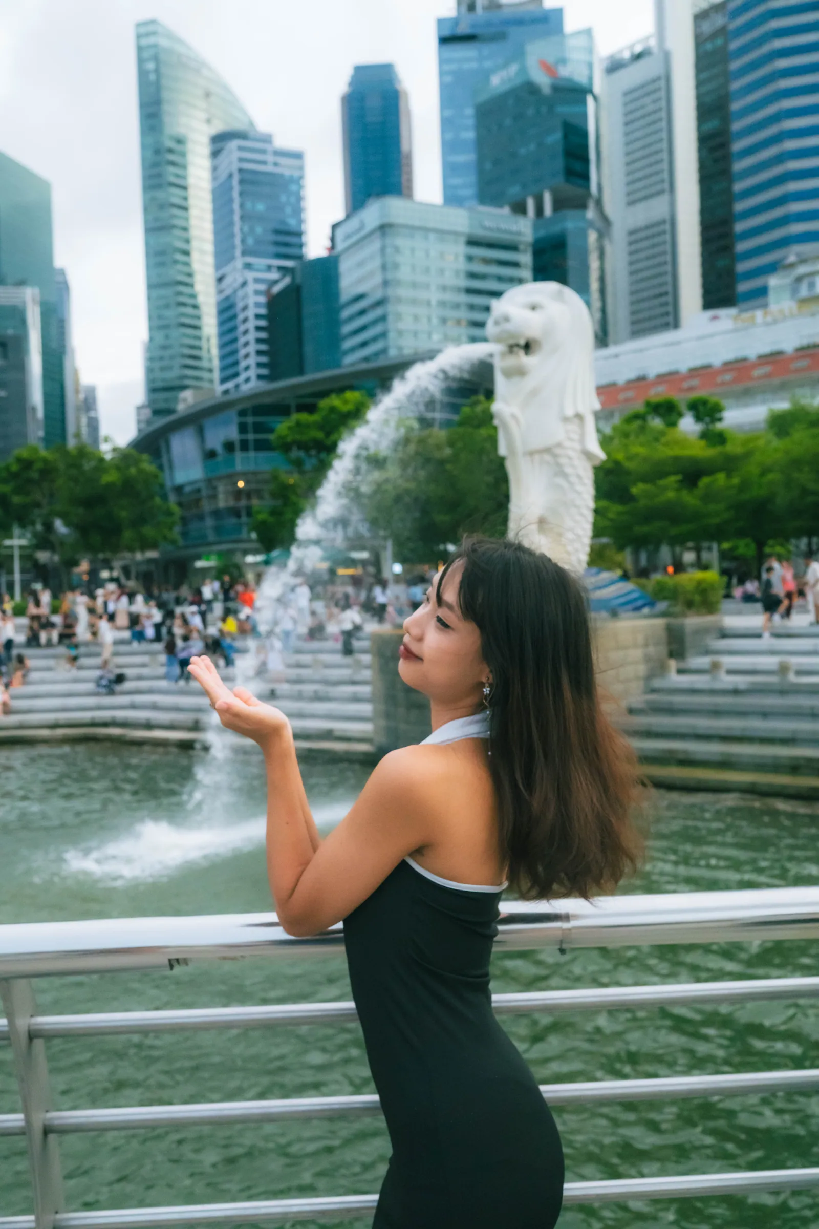 Woman looking back over her shoulder with her hand cupped as if catching the Merlion's water, skyline behind, Merlion Park