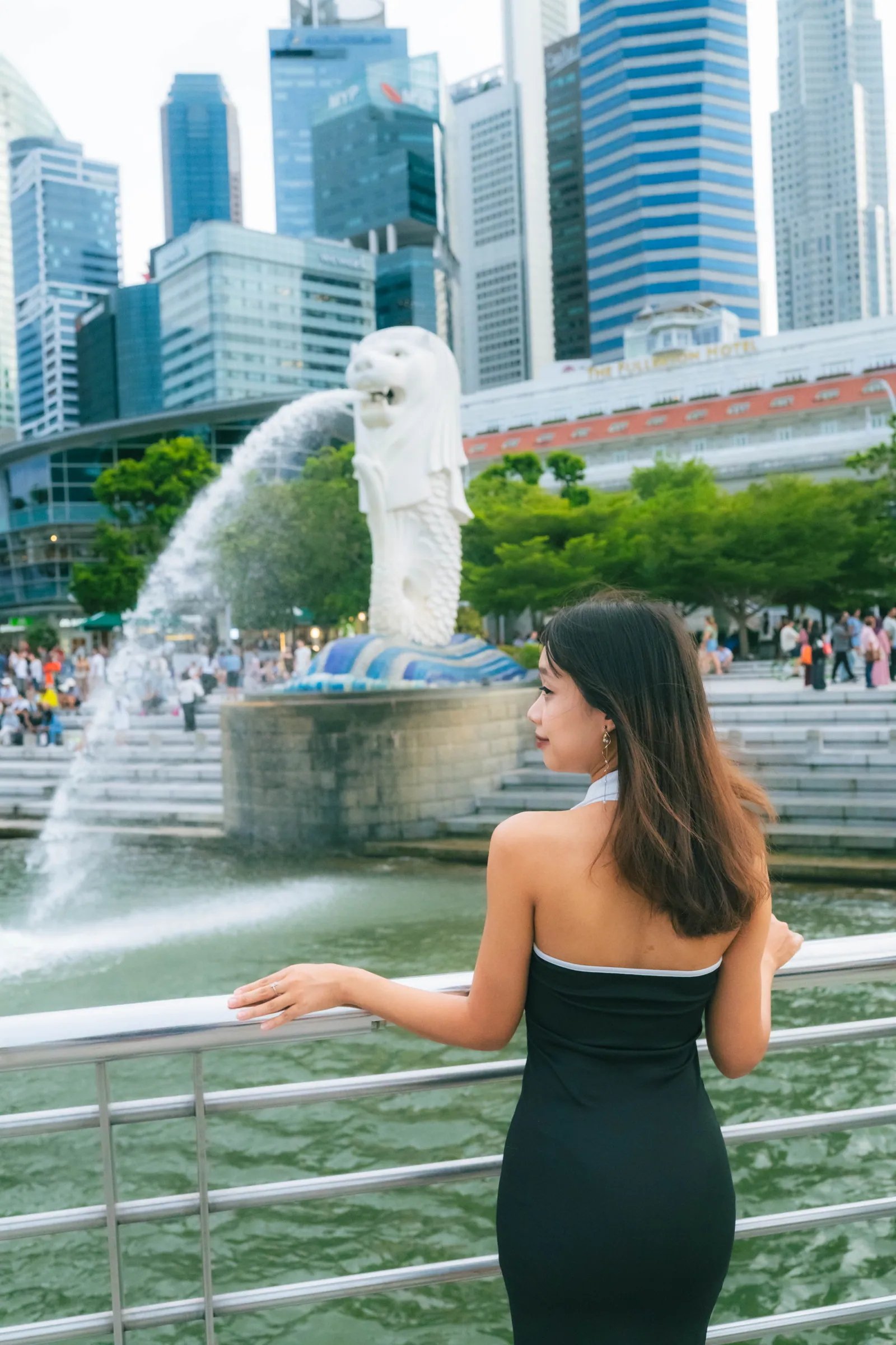 Woman in a black dress seen from behind, resting on the railing and looking at the Merlion, with The Fullerton Hotel behind, Merlion Park