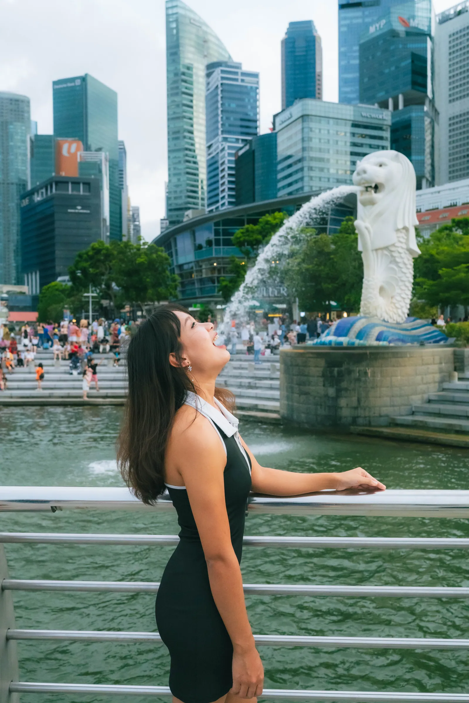 Woman in a black dress pretending to drink the water spouting from the Merlion statue, with the Singapore skyline behind, Merlion Park photoshoot