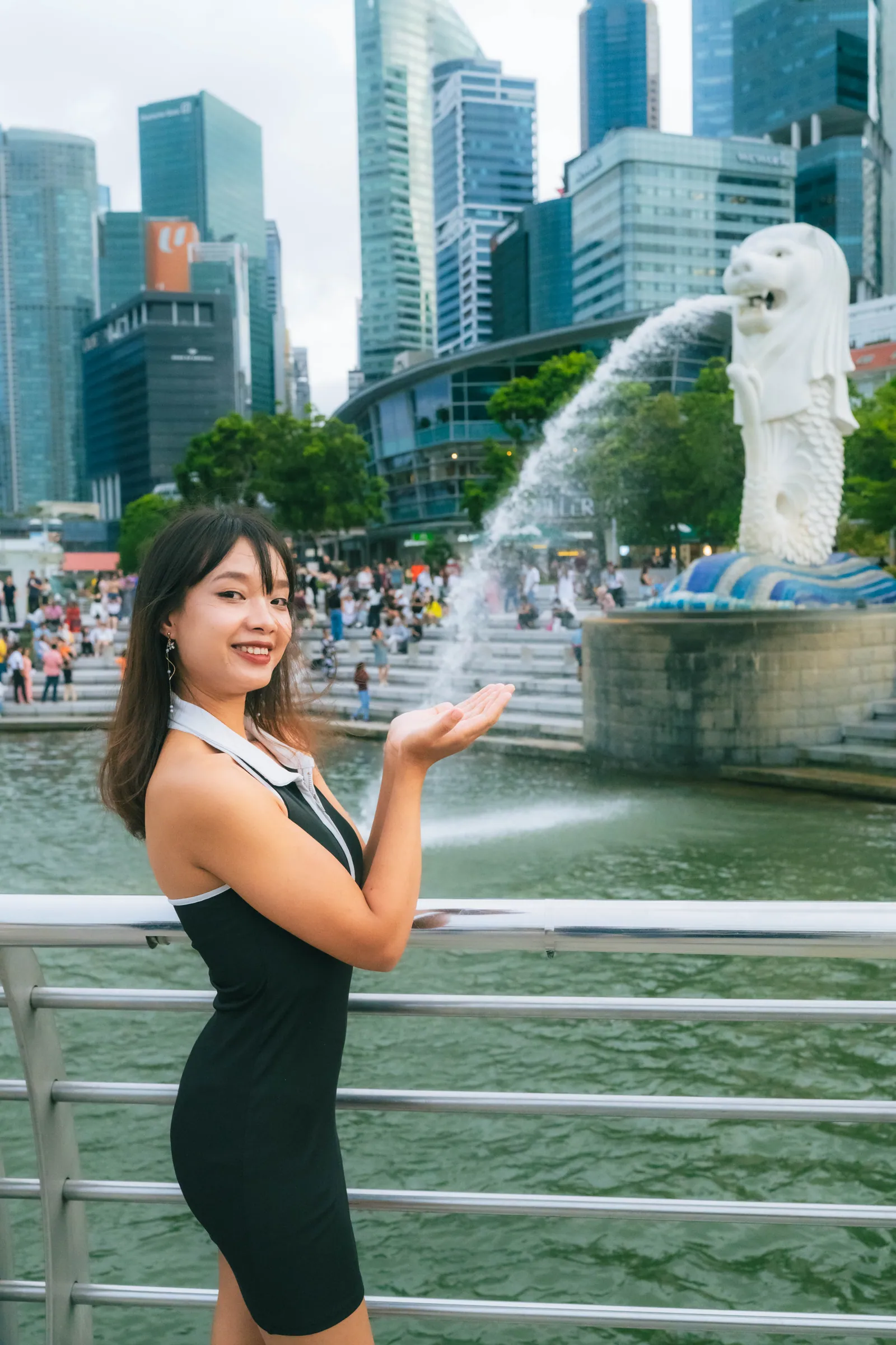 Woman in a black dress smiling and holding out her palm so the Merlion's water looks like it lands in her hand, Merlion Park, Singapore