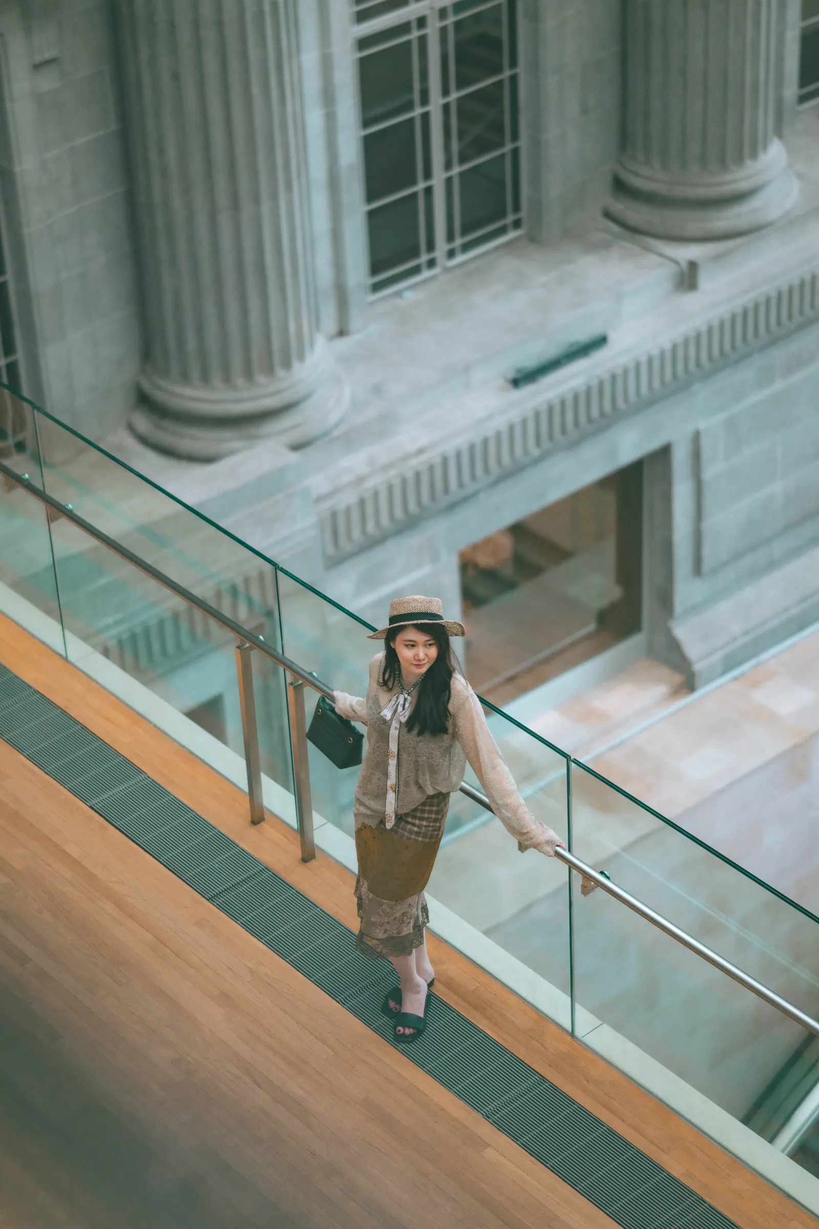 Woman in a hat walking down a sloping wooden walkway with a glass railing, beside tall stone columns, National Gallery Singapore