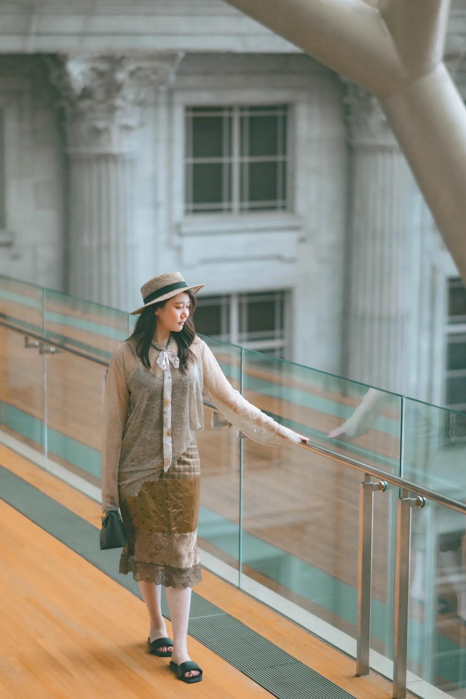 Woman in a hat resting her hand on a glass railing, with the stone facade and windows of the old Supreme Court behind, National Gallery Singapore