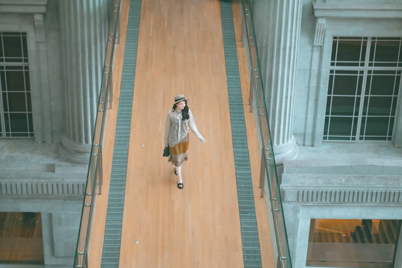Woman in a hat walking along a wooden linkbridge between the stone columns of the old Supreme Court and City Hall, National Gallery Singapore