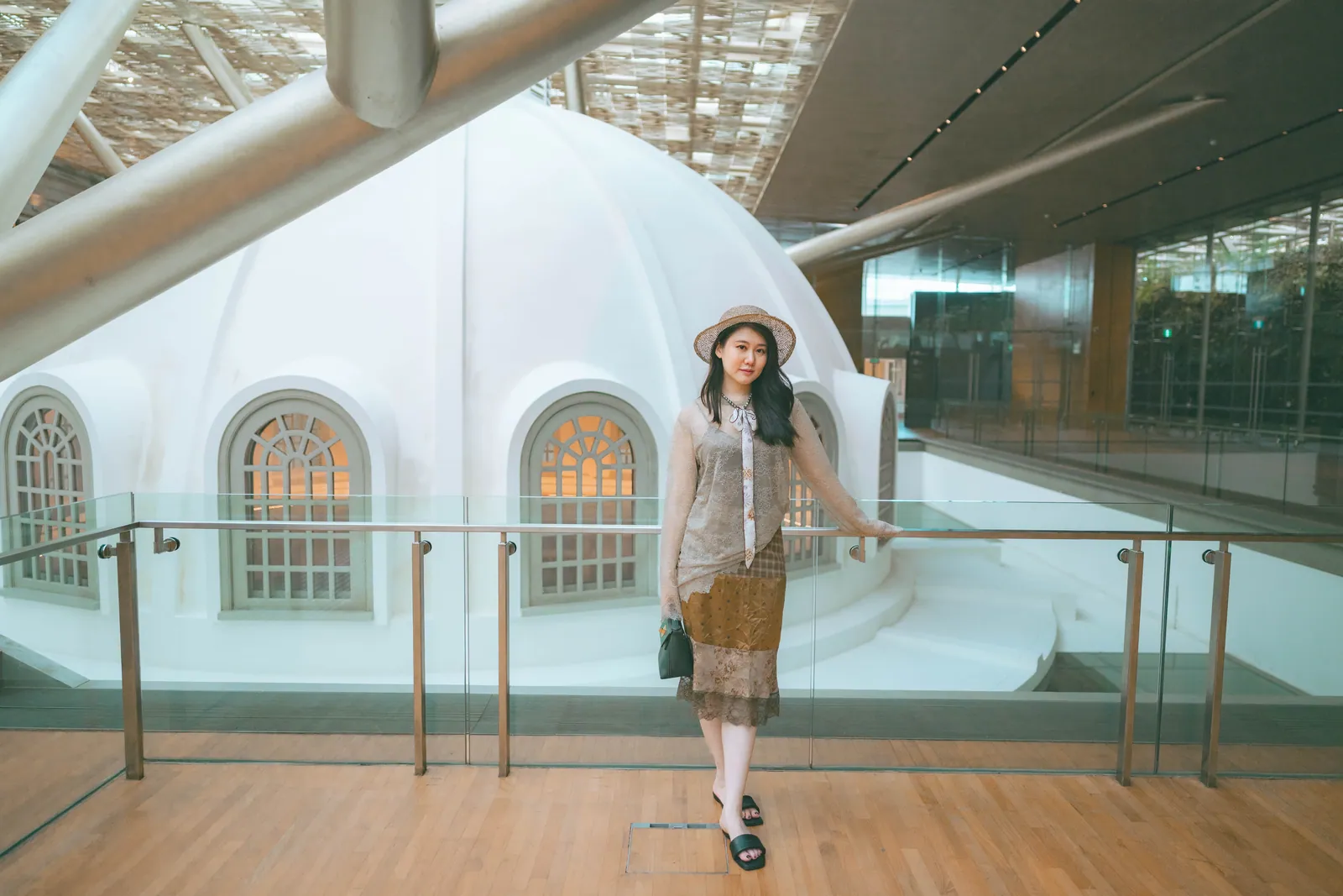 Woman in a hat and patterned dress standing on a wooden deck in front of the white Supreme Court dome under the glass roof, National Gallery Singapore
