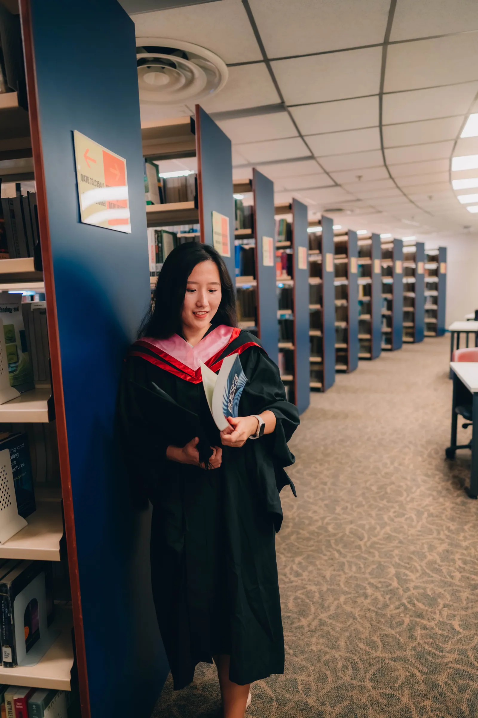 Master's graduate in gown leaning against a bookshelf and reading in the NTU library, Singapore