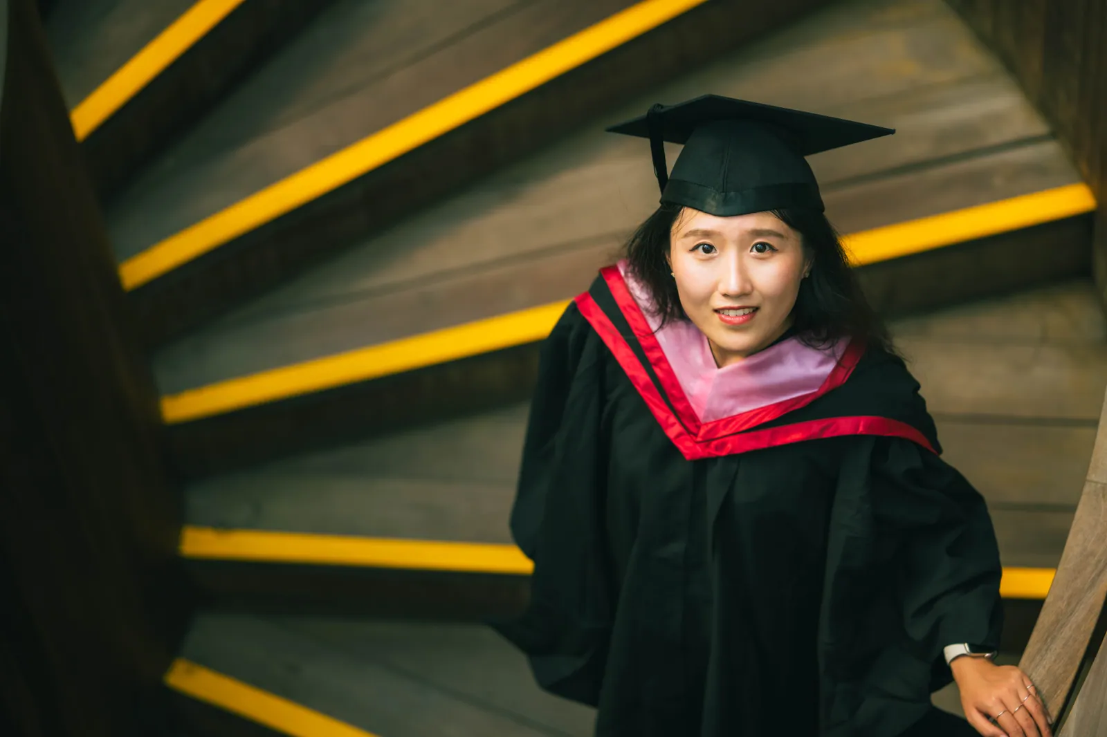 NTU master's graduate in cap and gown on a curving staircase with bright yellow step edges