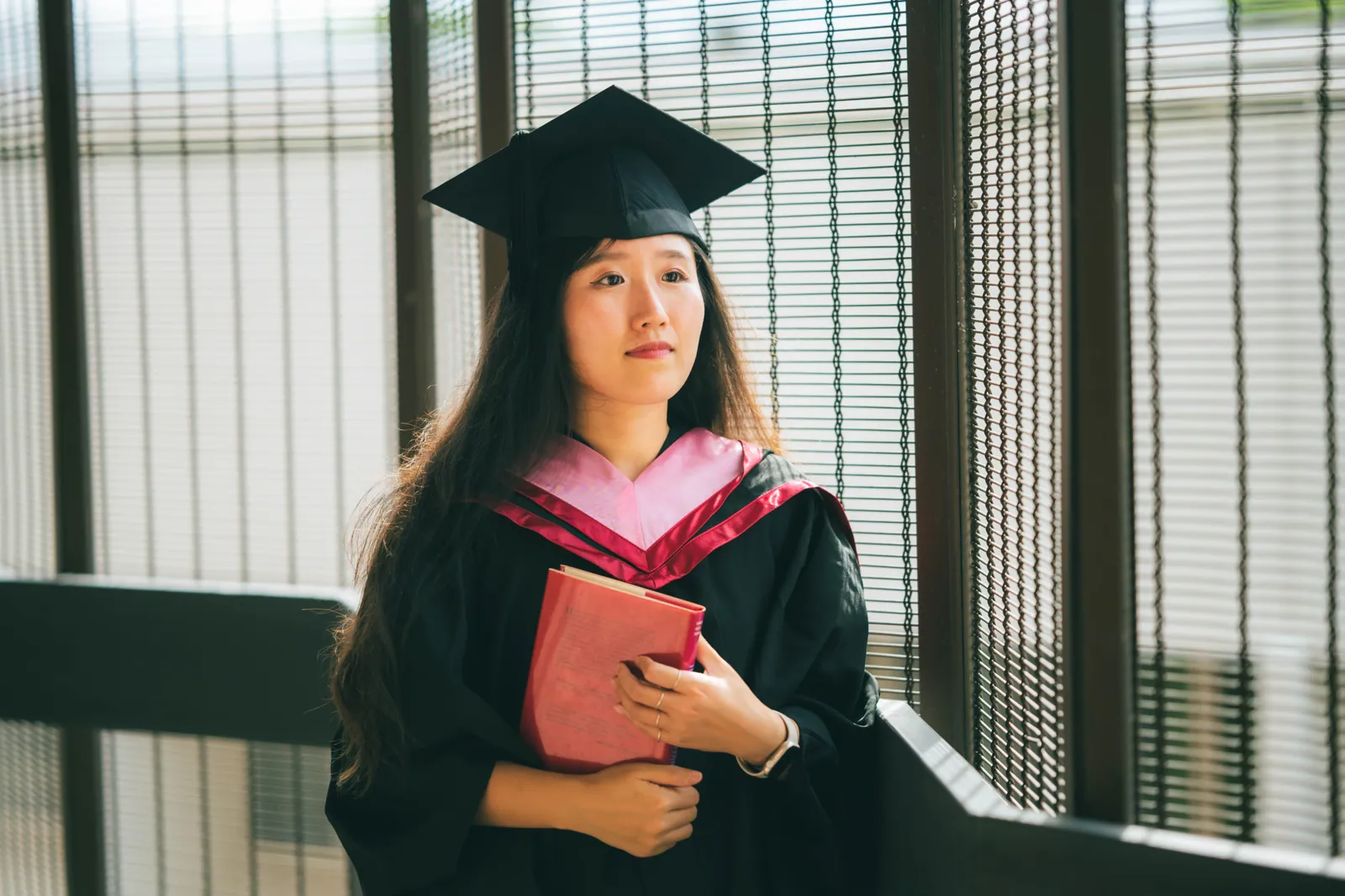 NTU graduate in cap and gown holding a red book by a window with soft light