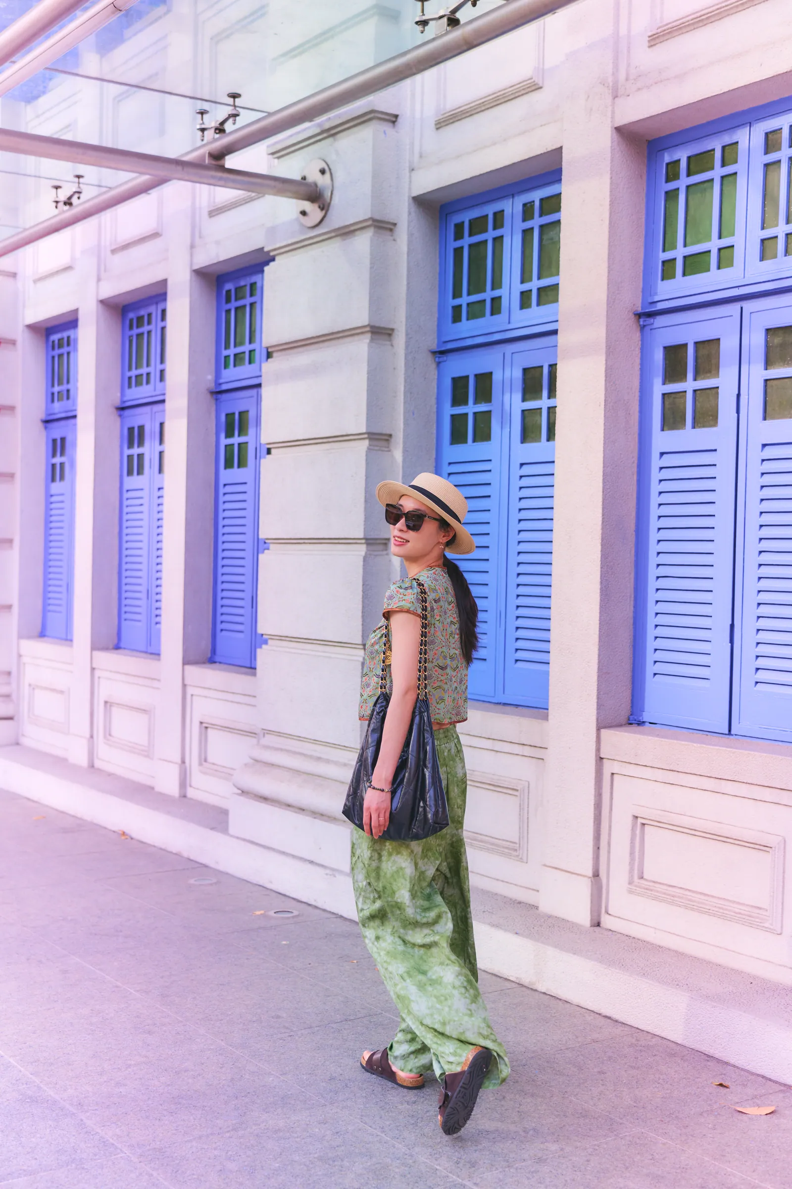 Woman in a hat and green trousers walking past tall blue wooden shutters and white walls, Old Hill Street Police Station
