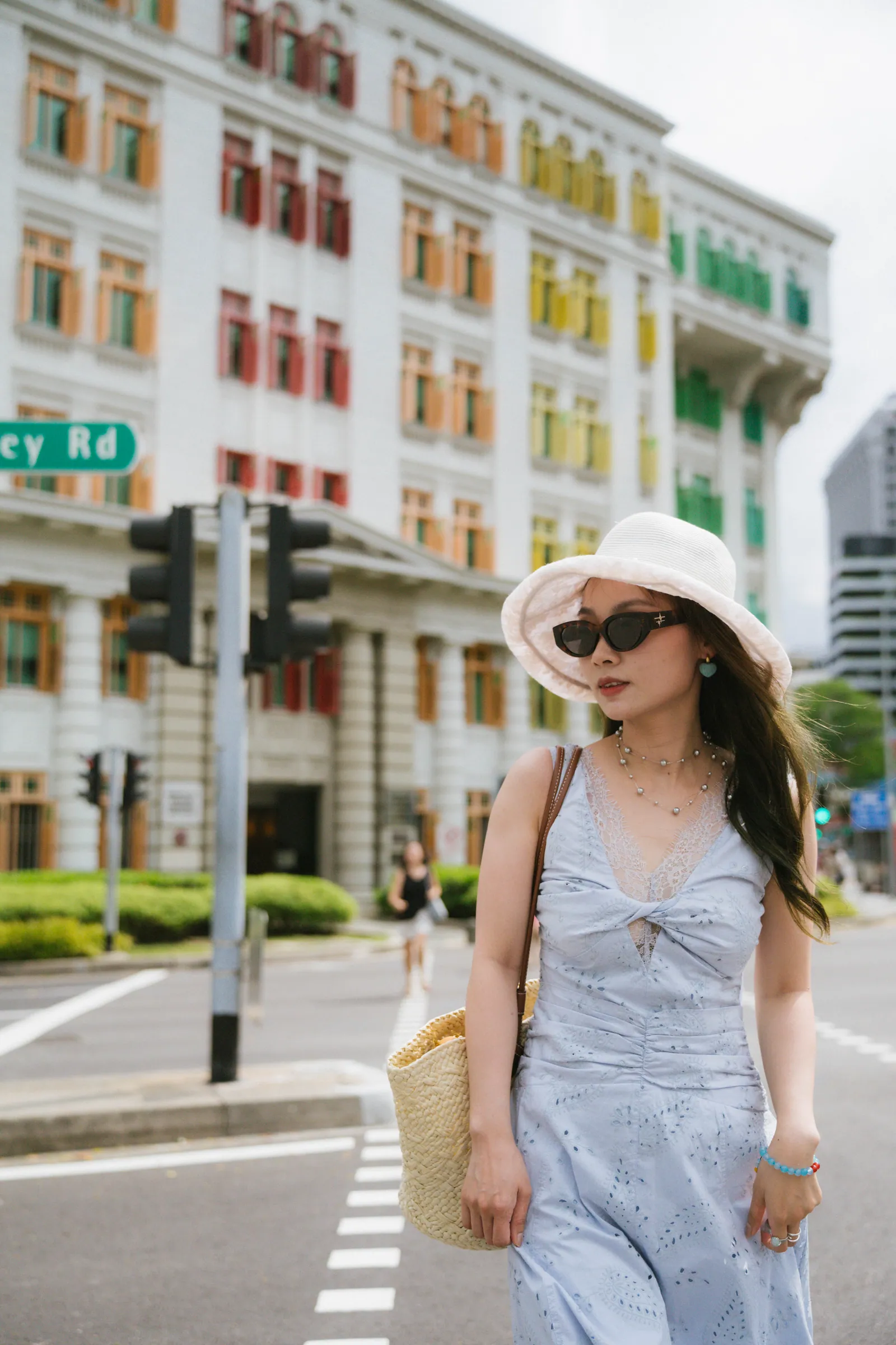 Woman in a light blue dress, sunglasses and white hat walking on a crossing, with the rainbow window facade behind her, Old Hill Street Police Station