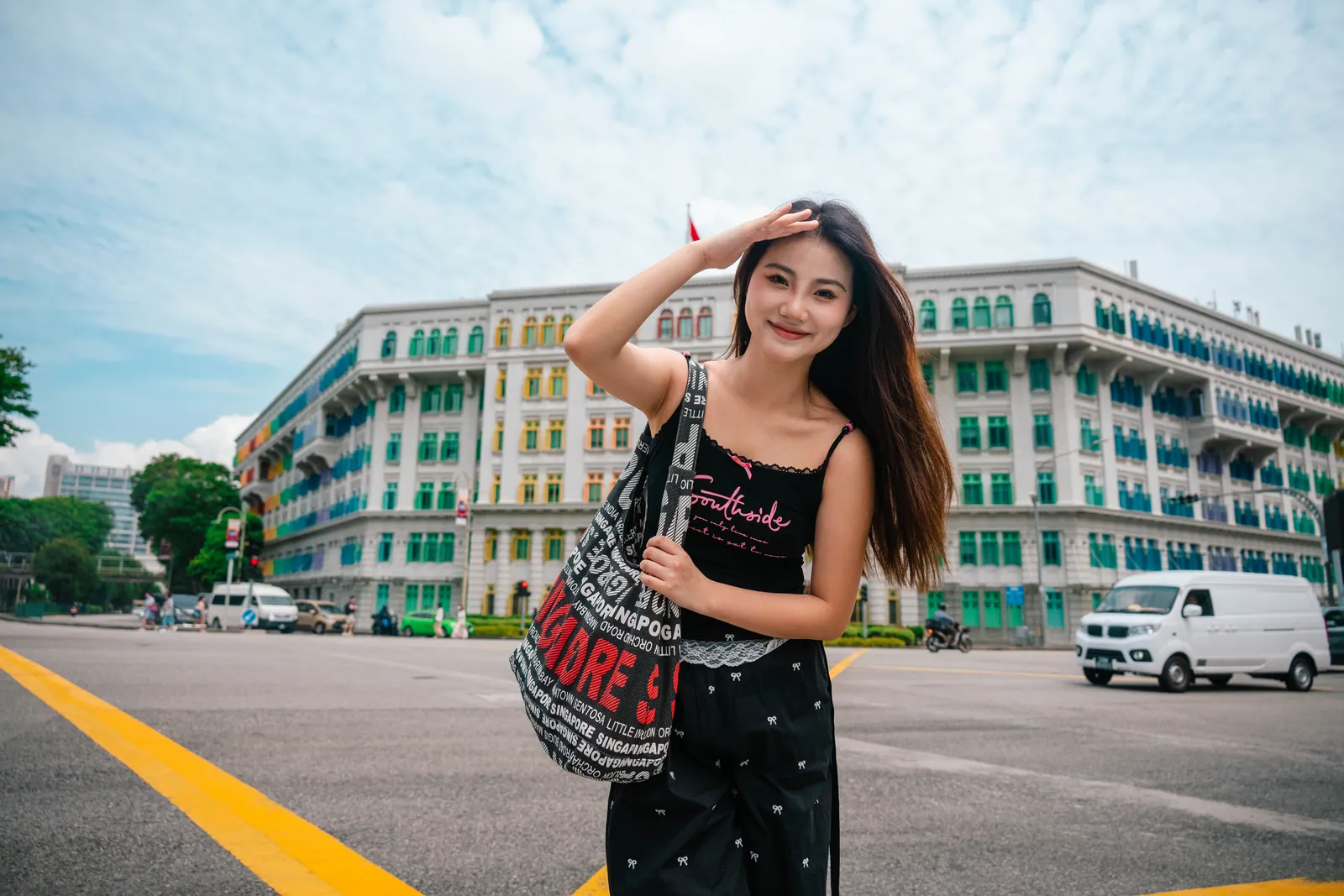 Woman in a black outfit smiling with a hand on her head, with the long rainbow-windowed facade of the Old Hill Street Police Station behind, Singapore