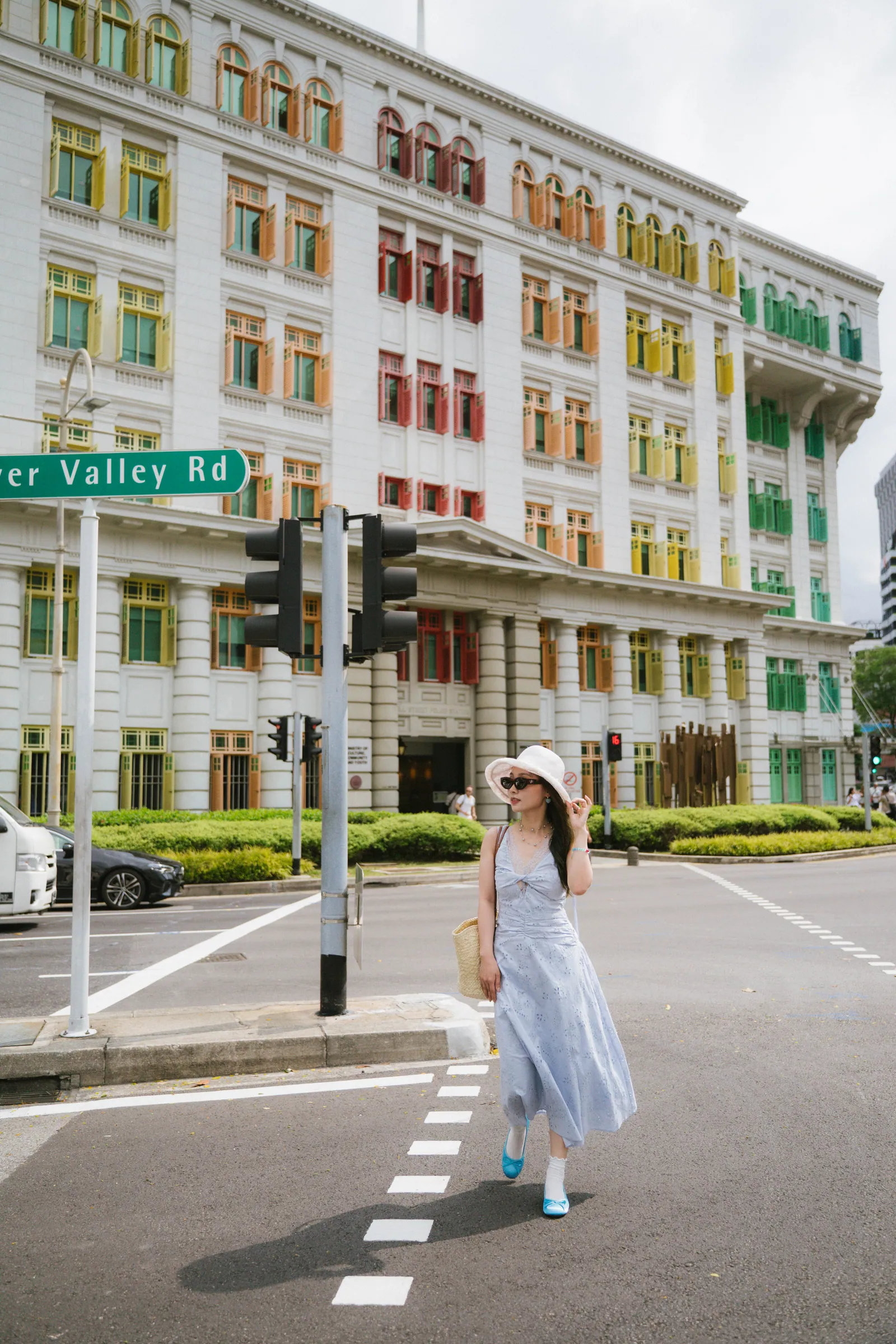 新加坡彩虹警察局旅拍:穿浅蓝色连衣裙、戴遮阳帽的女生在彩虹窗大楼前过马路