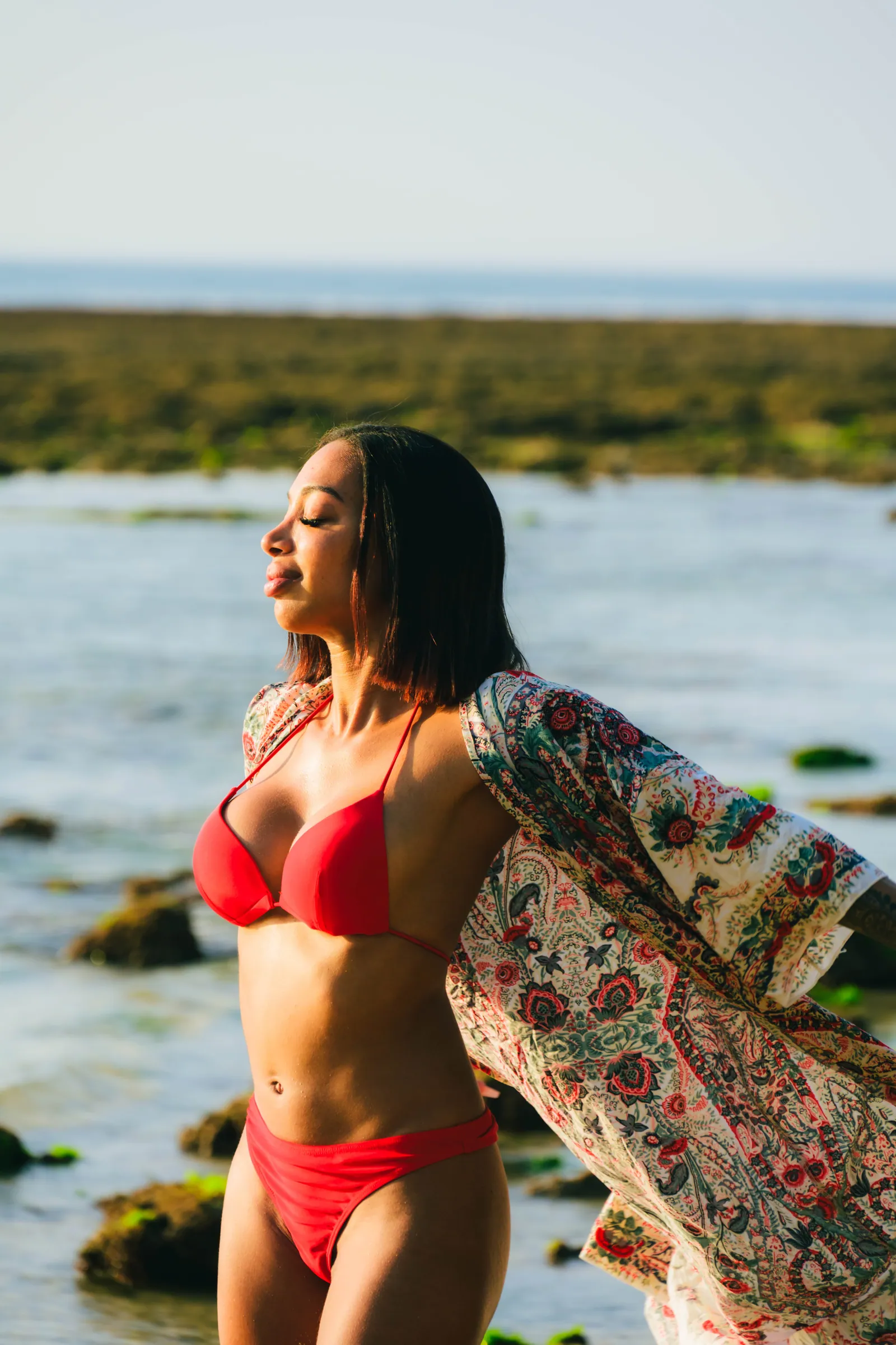 Woman in a red bikini and printed kimono with her eyes closed, facing the golden sunset light at Padang Padang Beach